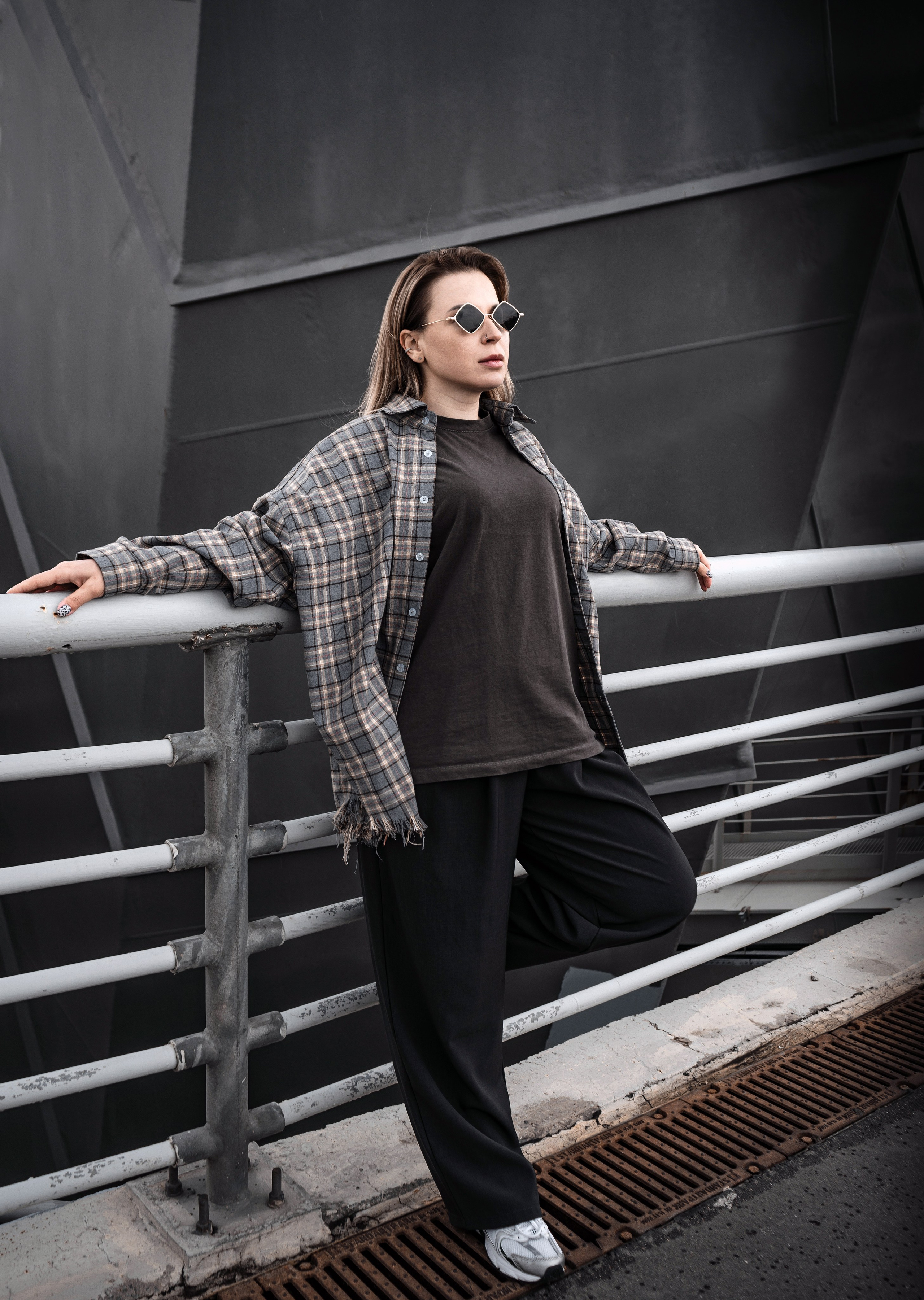 Urban street style портрет: девушка в oversize клетчатой рубашке и baggy чёрных брюках опирается на перила моста, развевающиеся волосы, тёмные cat-eye очки, индустриальный городской бэкграунд, ветреная погода