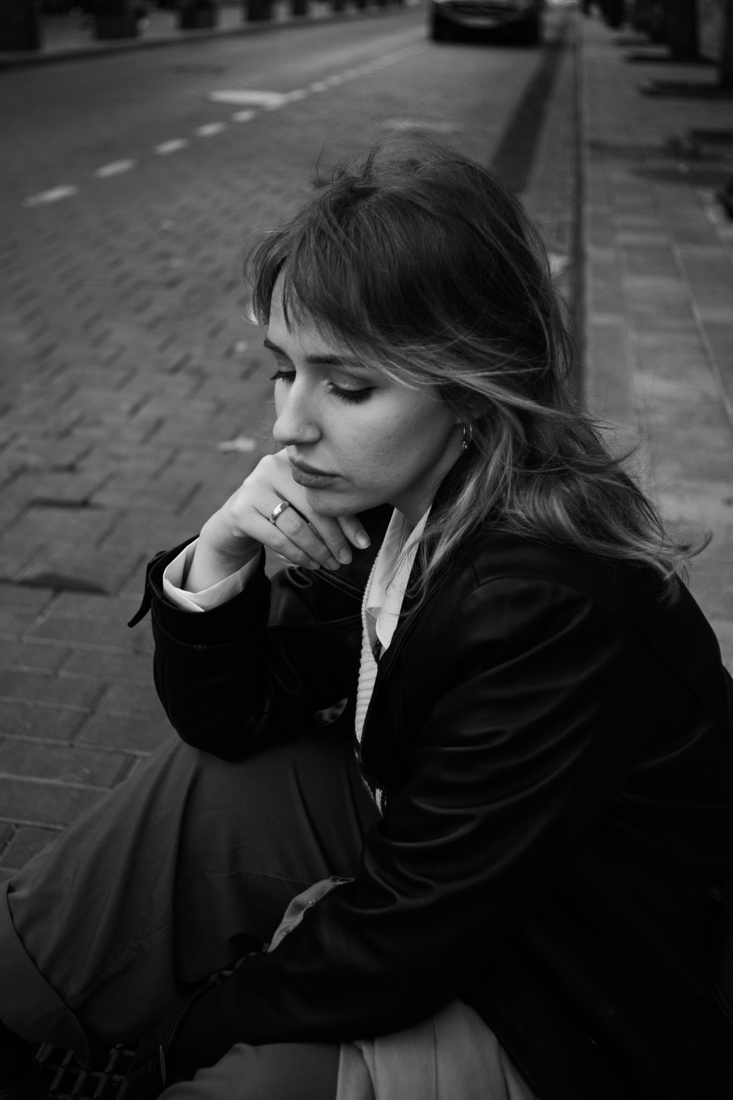 Чёрно-белый портрет женщины, сидящей на городской брусчатке с задумчивым выражением лица, в кожаной куртке и с развевающимися волосами, взгляд опущен вниз.