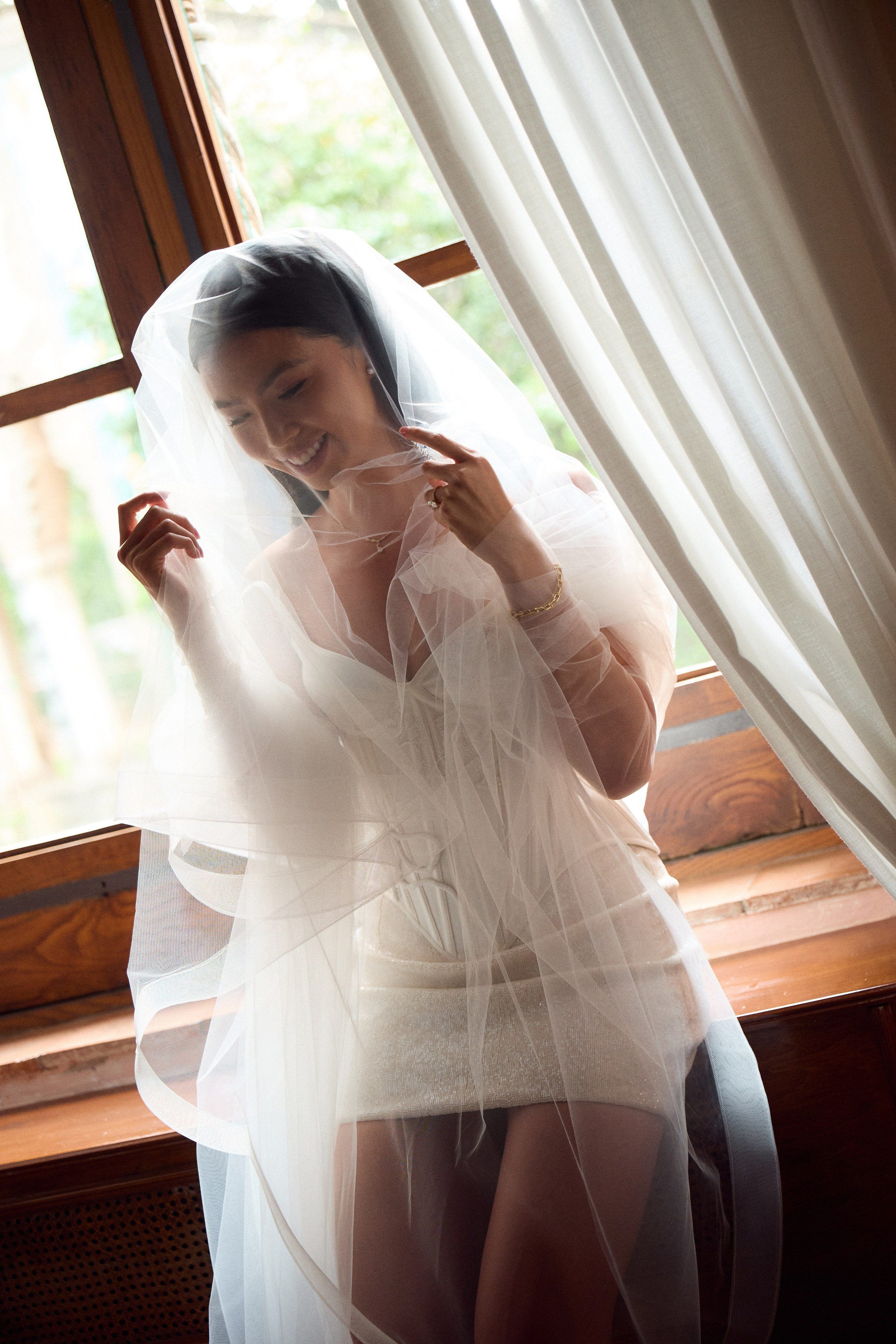 Bride adjusting her veil beside a sunlit window 