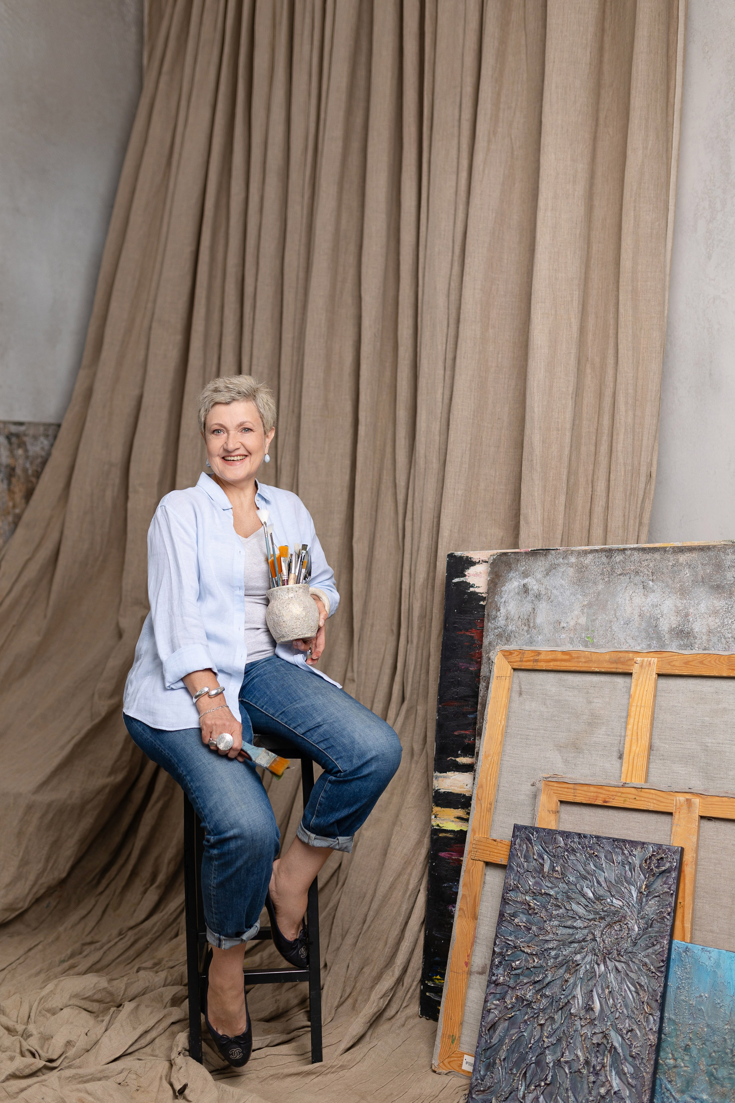 Художница держит в левой руке вазу с кистями,перед ней у стены стоят холсты.