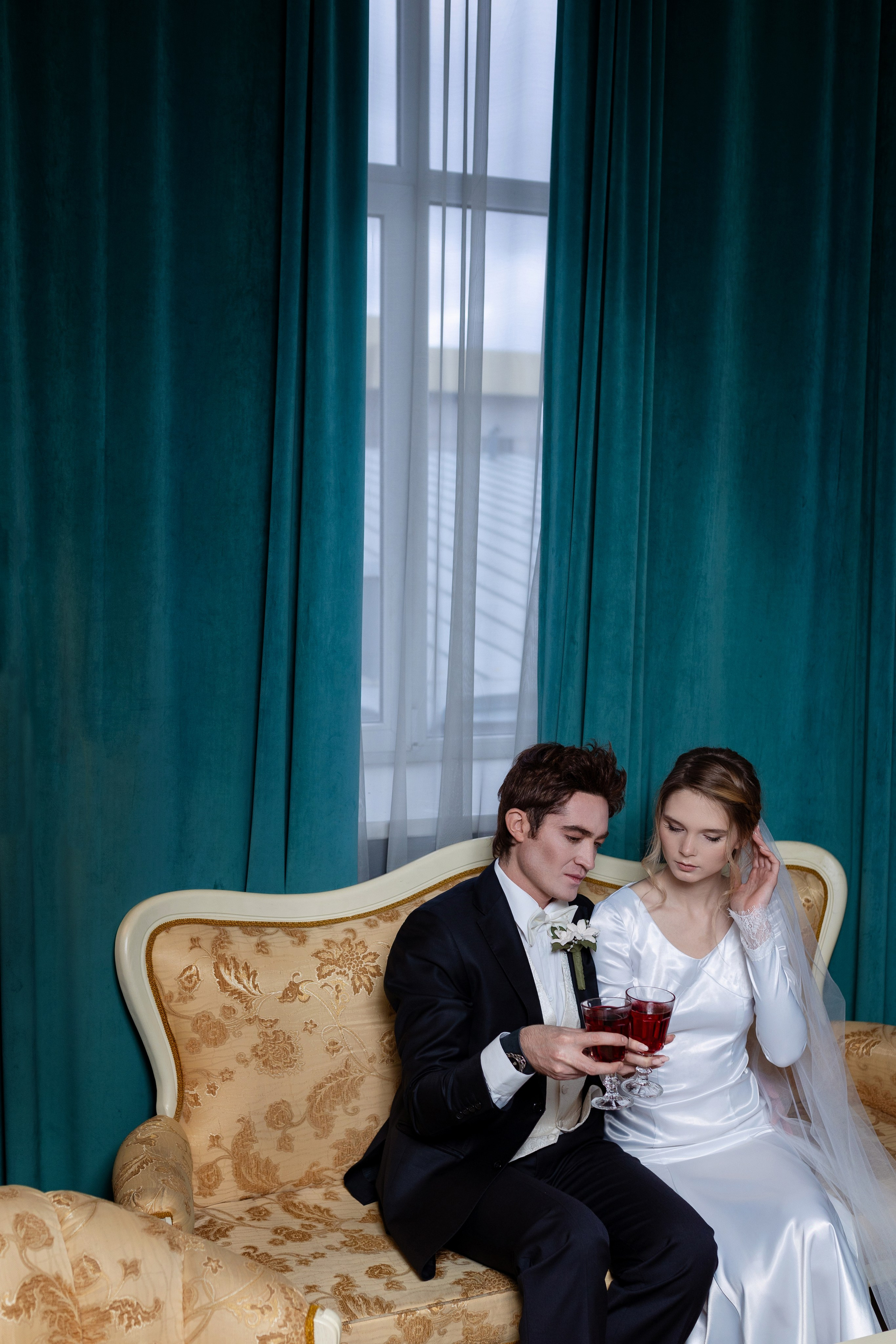 Жених и невеста сидят на диване на фоне окна.