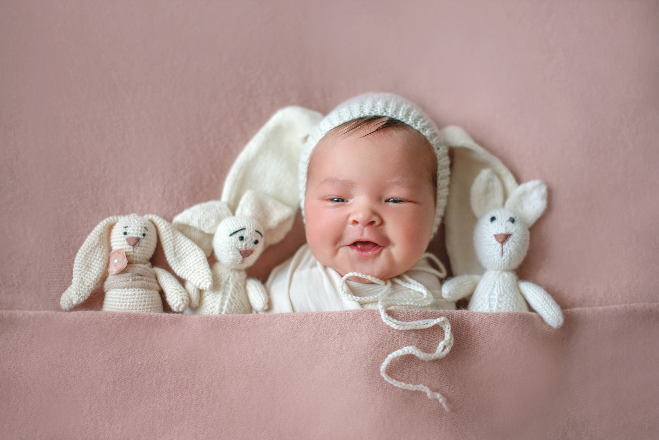 новорожденный лежит вместе с игрушками в шапочке зайчика и улыбается 