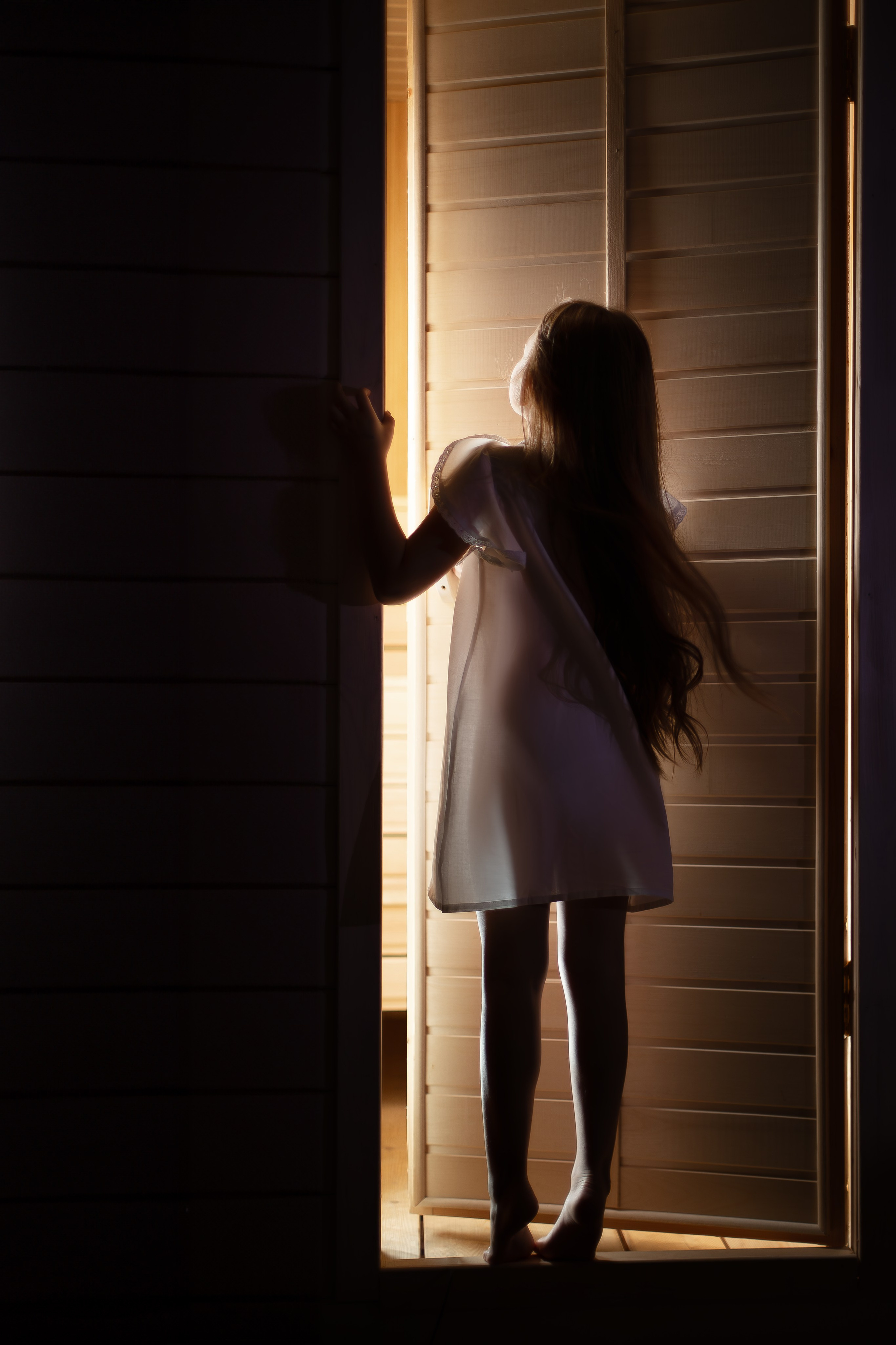 Девочка в светлом платье стоит в дверях, освещённая тёплым светом изнутри.