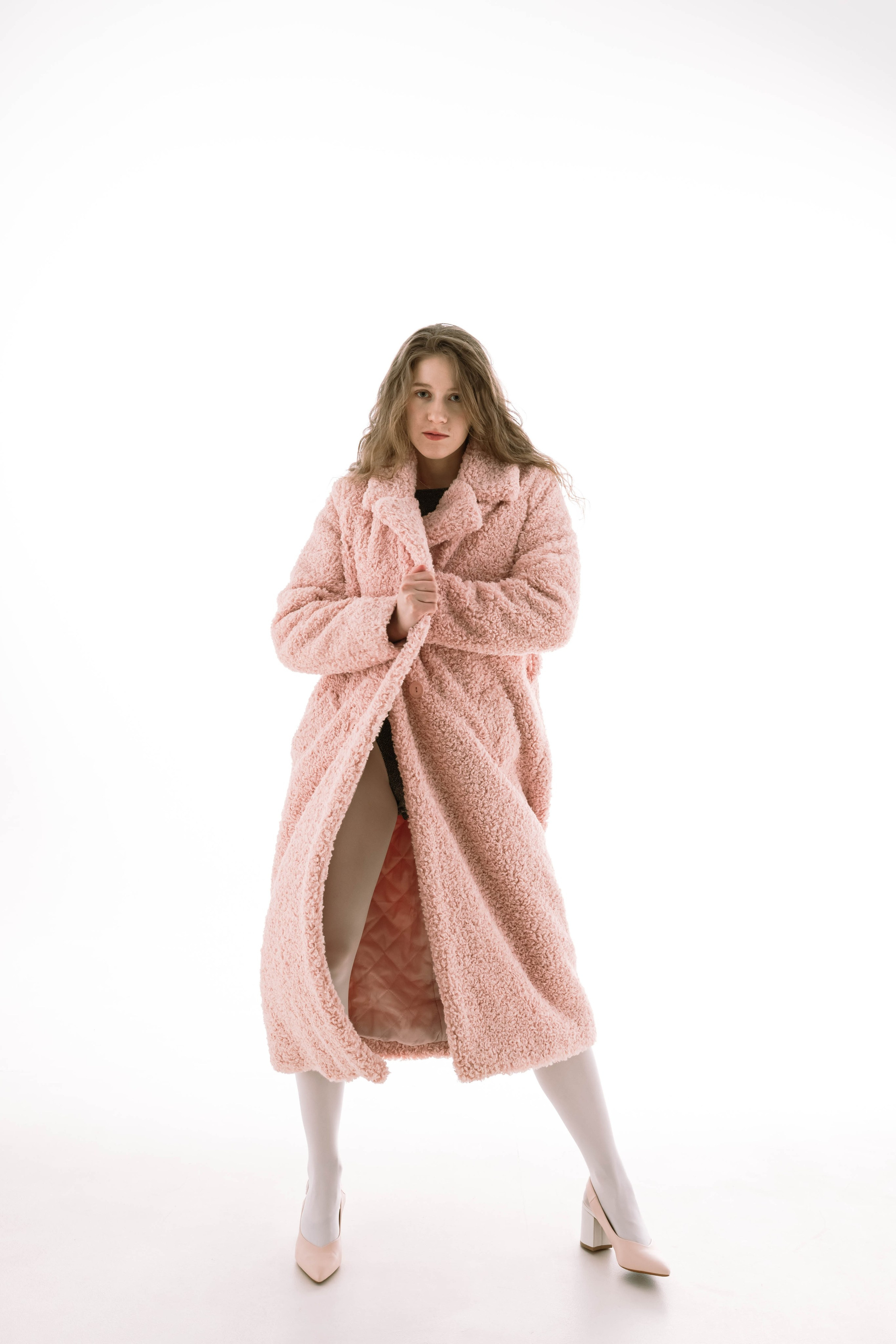 Модель смело смотрится в пышном розовом пальто и стильных туфлях на каблуках, демонстрируя модный дизайн на минималистичном белом фоне, излучая уверенность и элегантность.