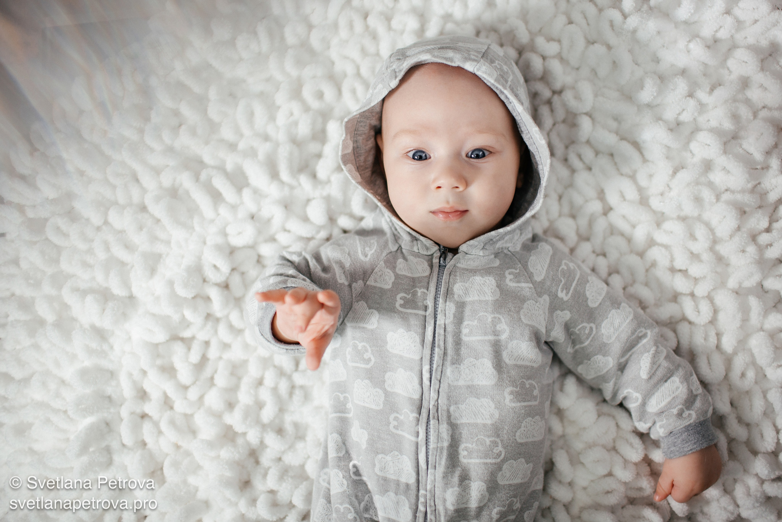 На пушистом белом ковре — взгляд в камеру. Любопытный малыш в сером комбинезоне с облачками. Малыш на мягком покрывале, спокойный взгляд. Пушистый фон, младенец в капюшоне — уютный момент.  Малыш изучает мир — лежит на белом ковре. Серый комбинезон с облаками, взгляд малыша. Тёплая фотосессия: младенец на мягком фоне.