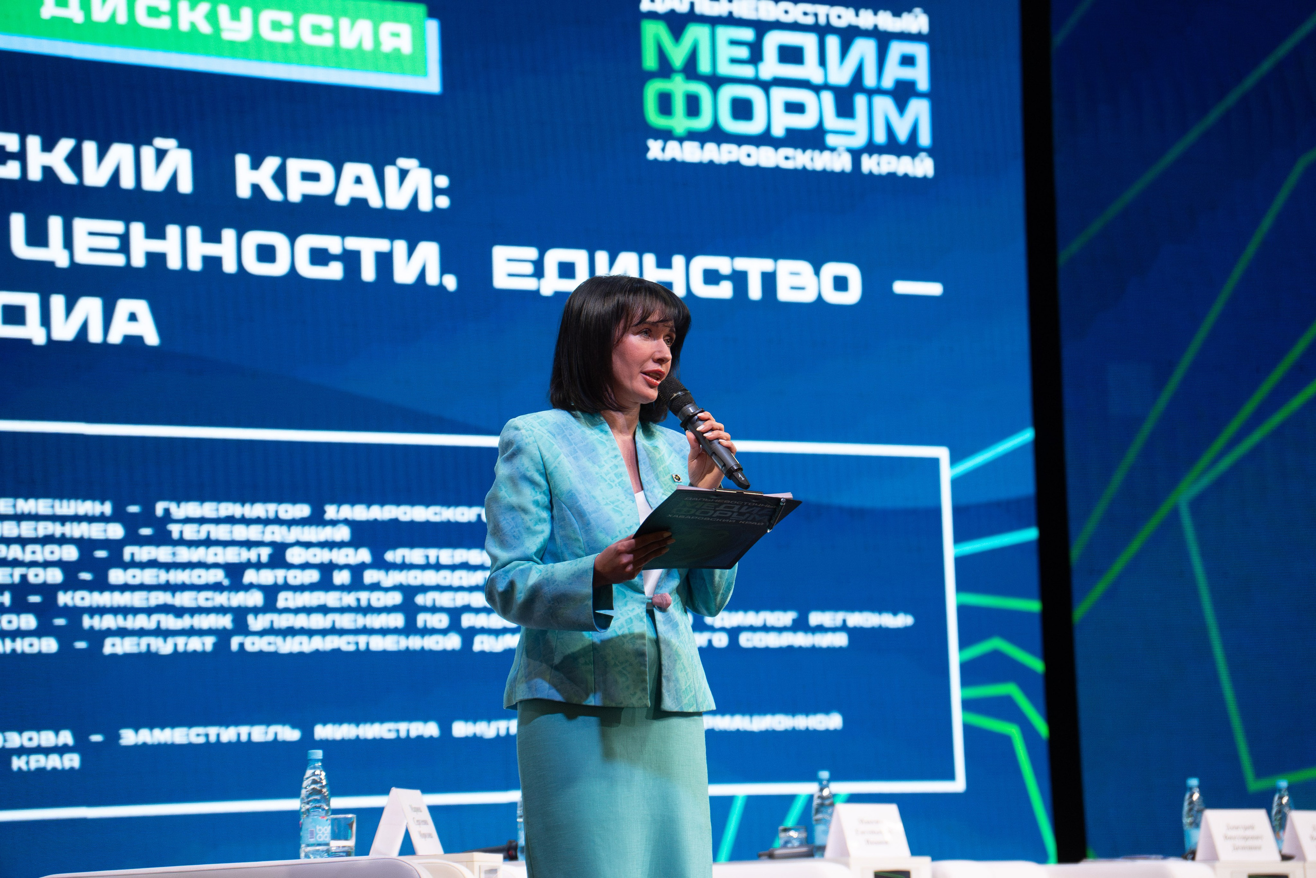 Репортажный фотограф в Хабаровске Дмитрий Дергачев снял ведущую мероприятия в ярком зеленом платье с микрофоном и папкой на сцене Дальневосточного медиафорума.
