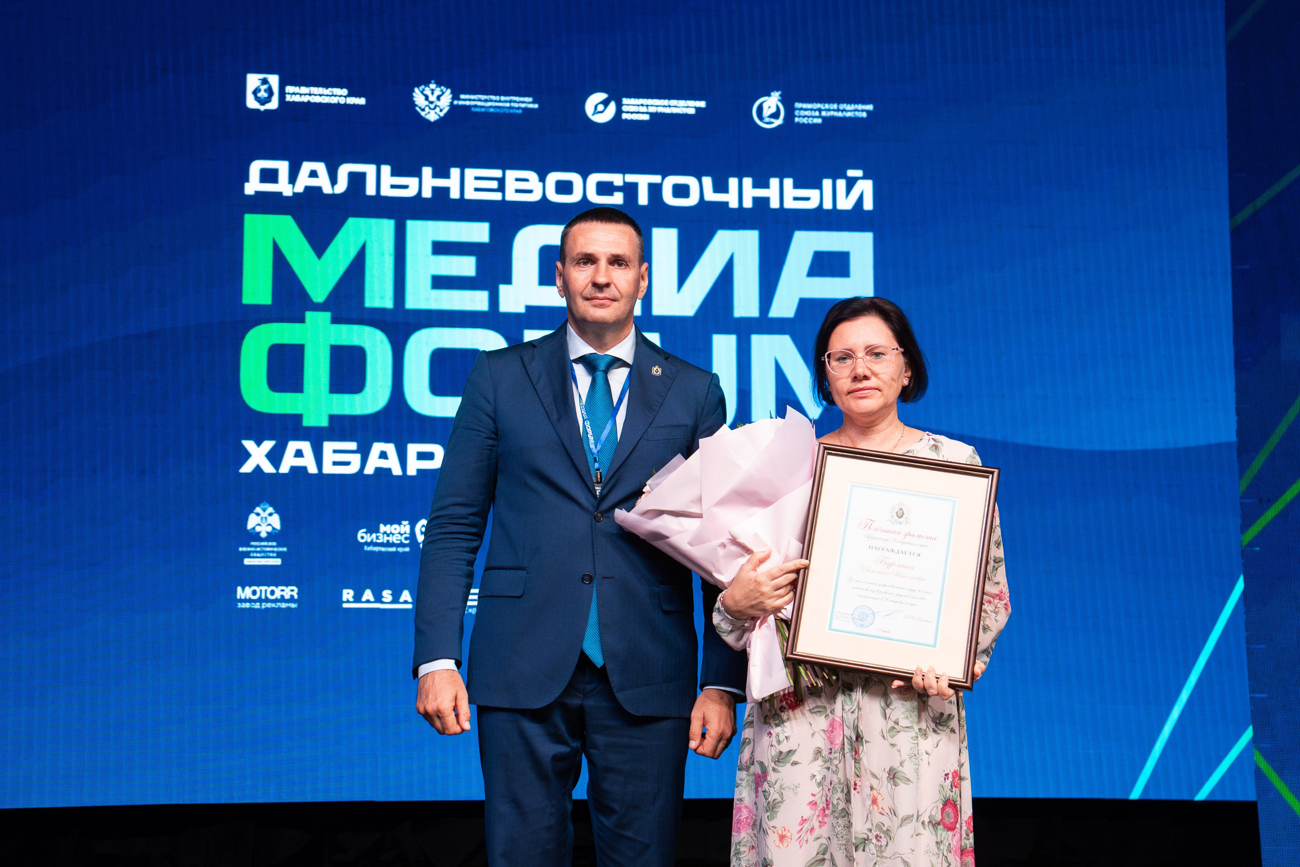 Репортажный фотограф в Хабаровске Дмитрий Дергачев заснял торжественное награждение женщины в цветочном платье с букетом и дипломом на сцене форума.