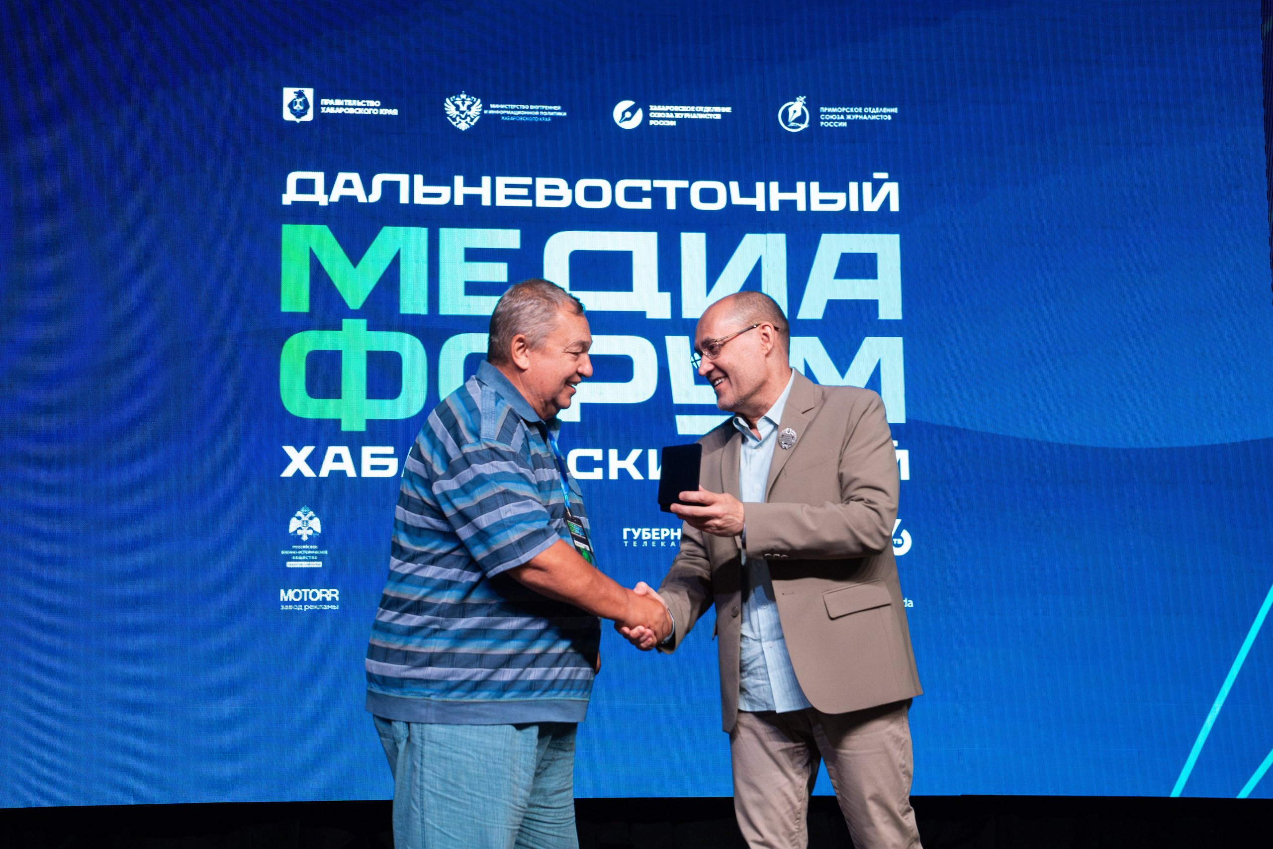 Кадр от репортажного фотографа в Хабаровске Дмитрия Дергачева: мужчины пожимают руки и передают награду на сцене Дальневосточного медиафорума.