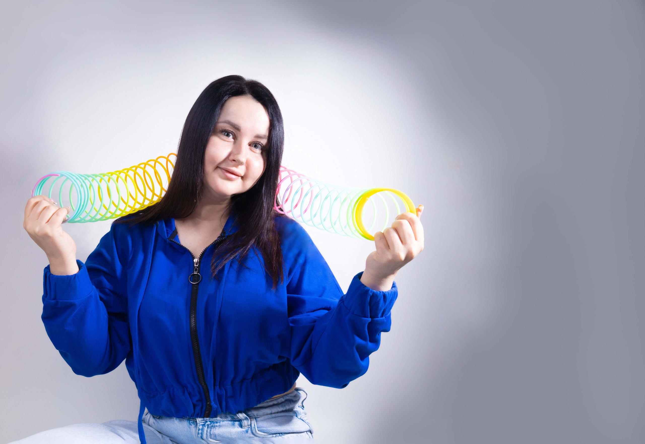 Молодая женщина в синей куртке держит разноцветную пружинку-игрушку (радужную спираль) обеими руками. Фон нейтральный, светлый.