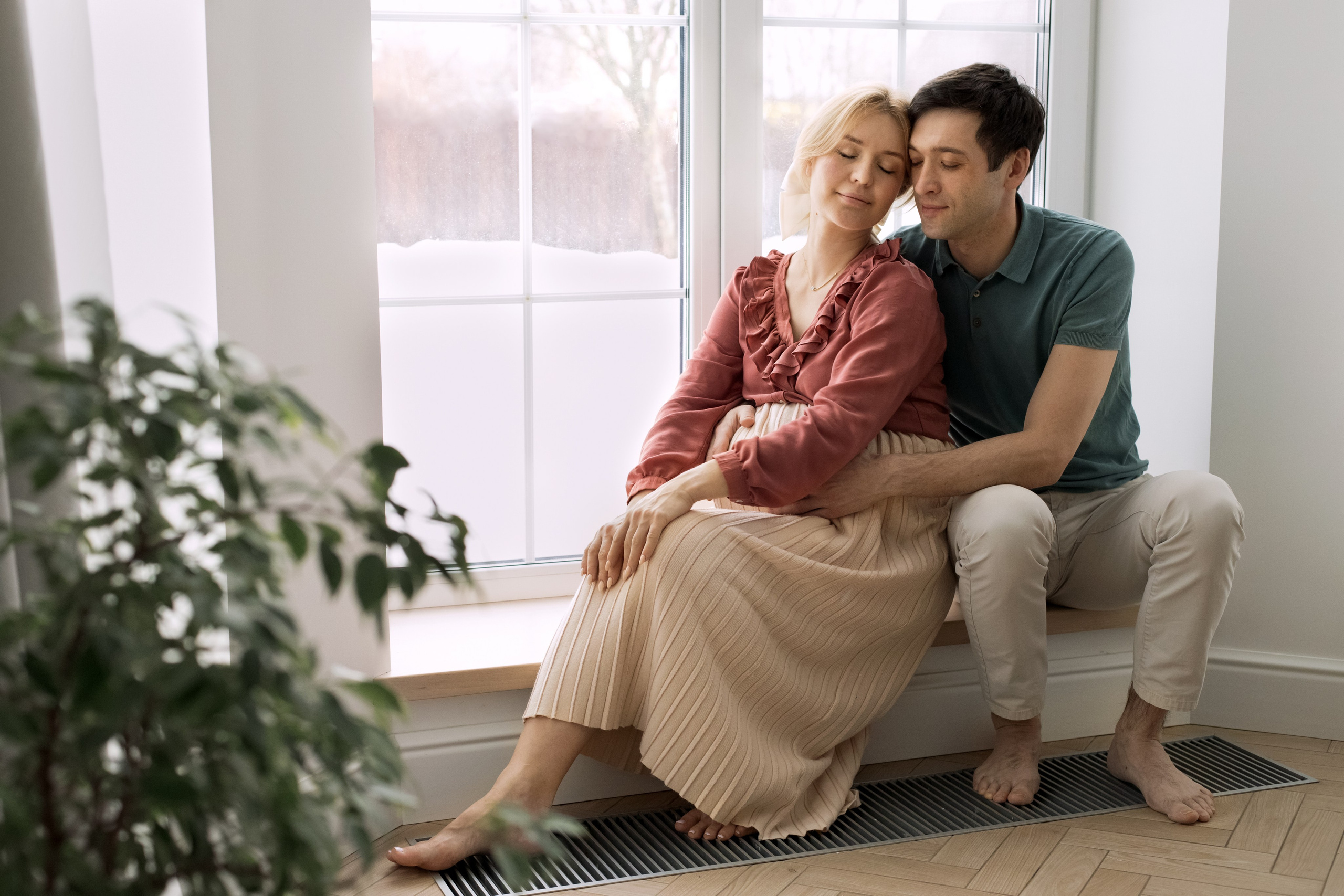 Муж обнимает беременную жену, сидя на подоконнике у большого окна
