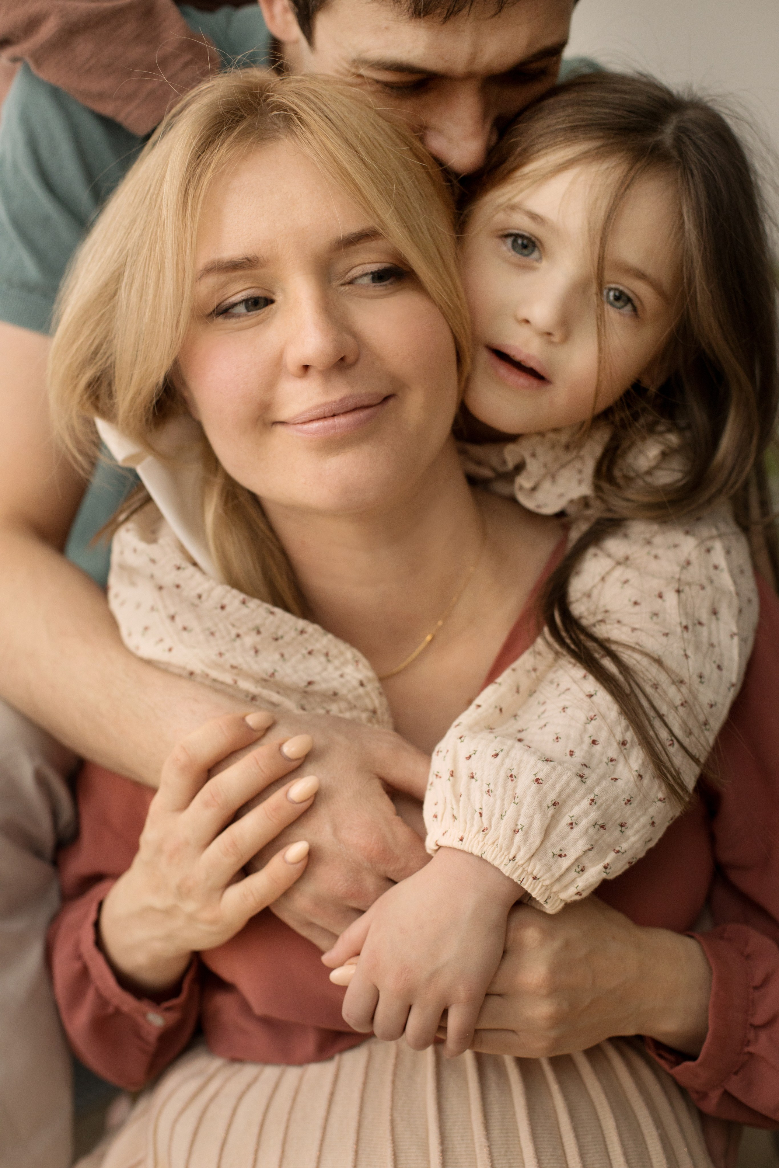 Девочка и папа обнимают беременную улыбающуюся маму со спины.