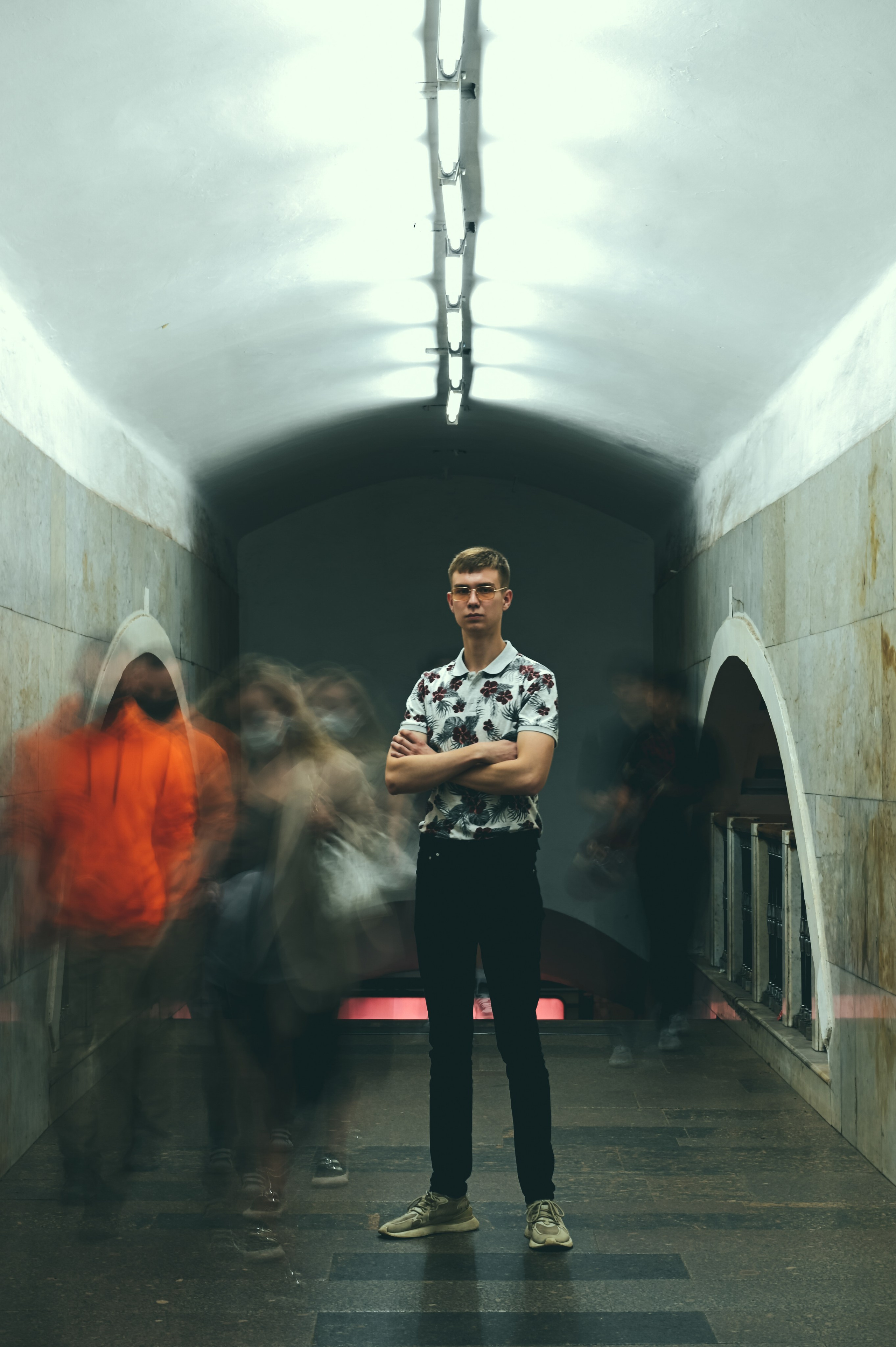 Молодой человек в стильной одежде стоит на фоне станции метро.  Освещение станции подчеркивает современный дизайн интерьера с арочными элементами и светильниками. Фотография передает динамику городской жизни и контраст между спокойствием модели и движением вокруг нее.