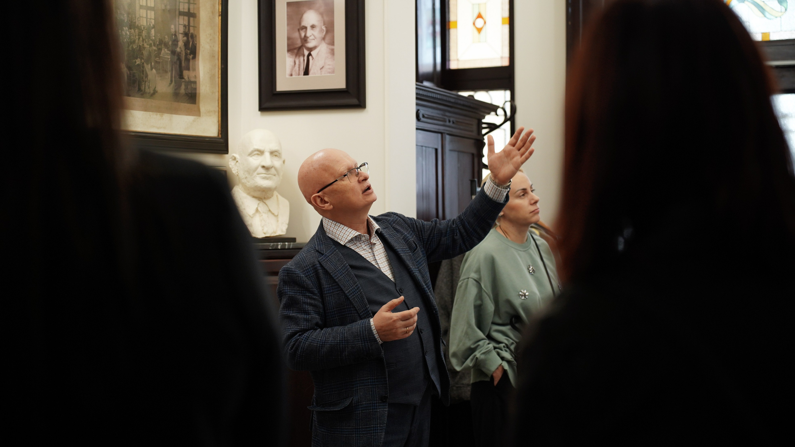 Мужчина Гордон  в Сочи проводит экскурсию по музею. Он стоит перед группой людей и указывает на экспонаты. Экспонатами являются бюст мужчины, картина с изображением мужчины и витраж. Мужчина одет в костюм и очки. Он лысый и с бородой. Люди в группе смотрят на экспонаты и слушают голос человека讲解.