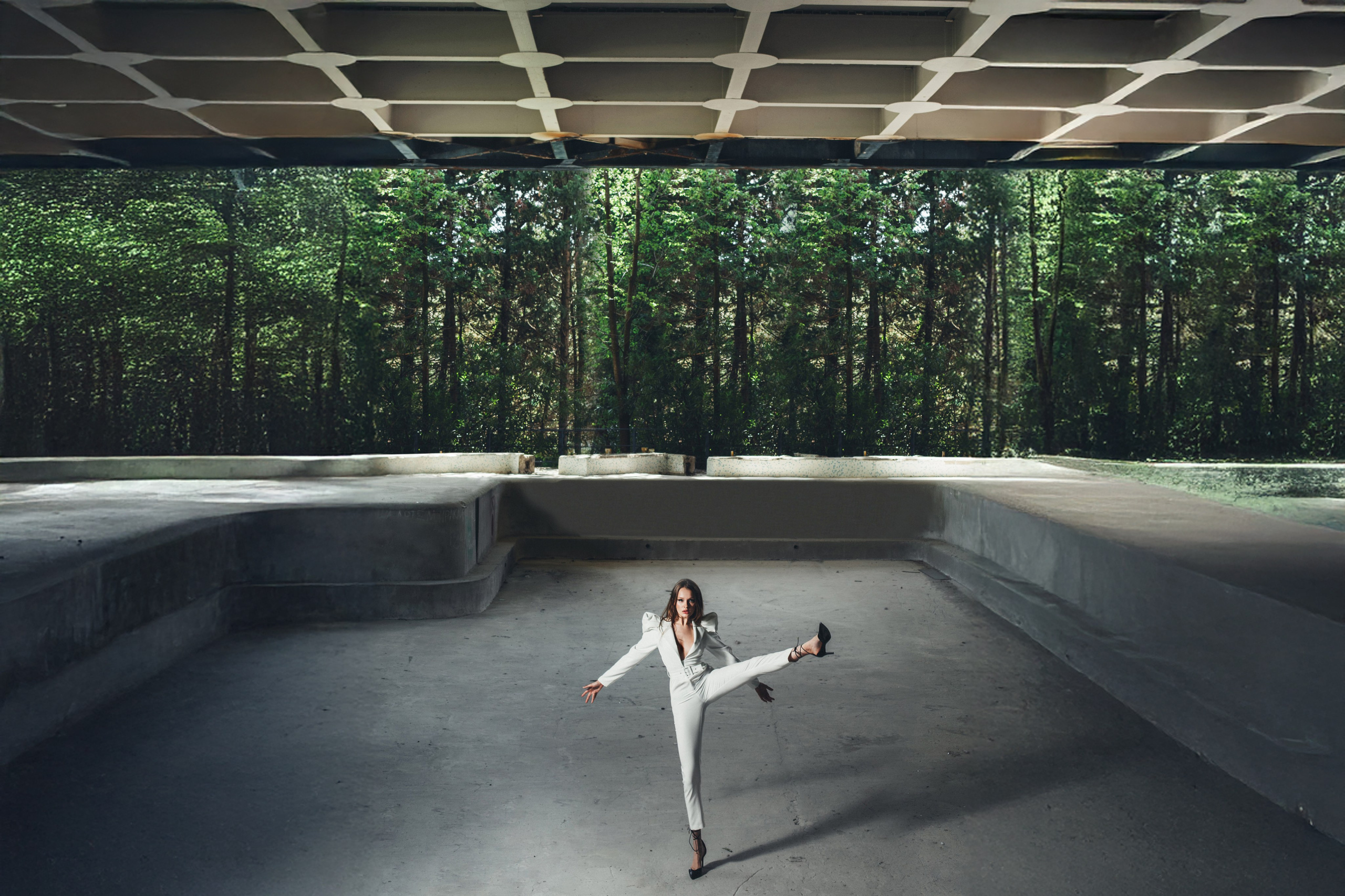 Девушка в белом костюме позирует в динамичной позе на фоне леса и геометрической бетонной конструкции — портретный фотограф Галина Скорик.