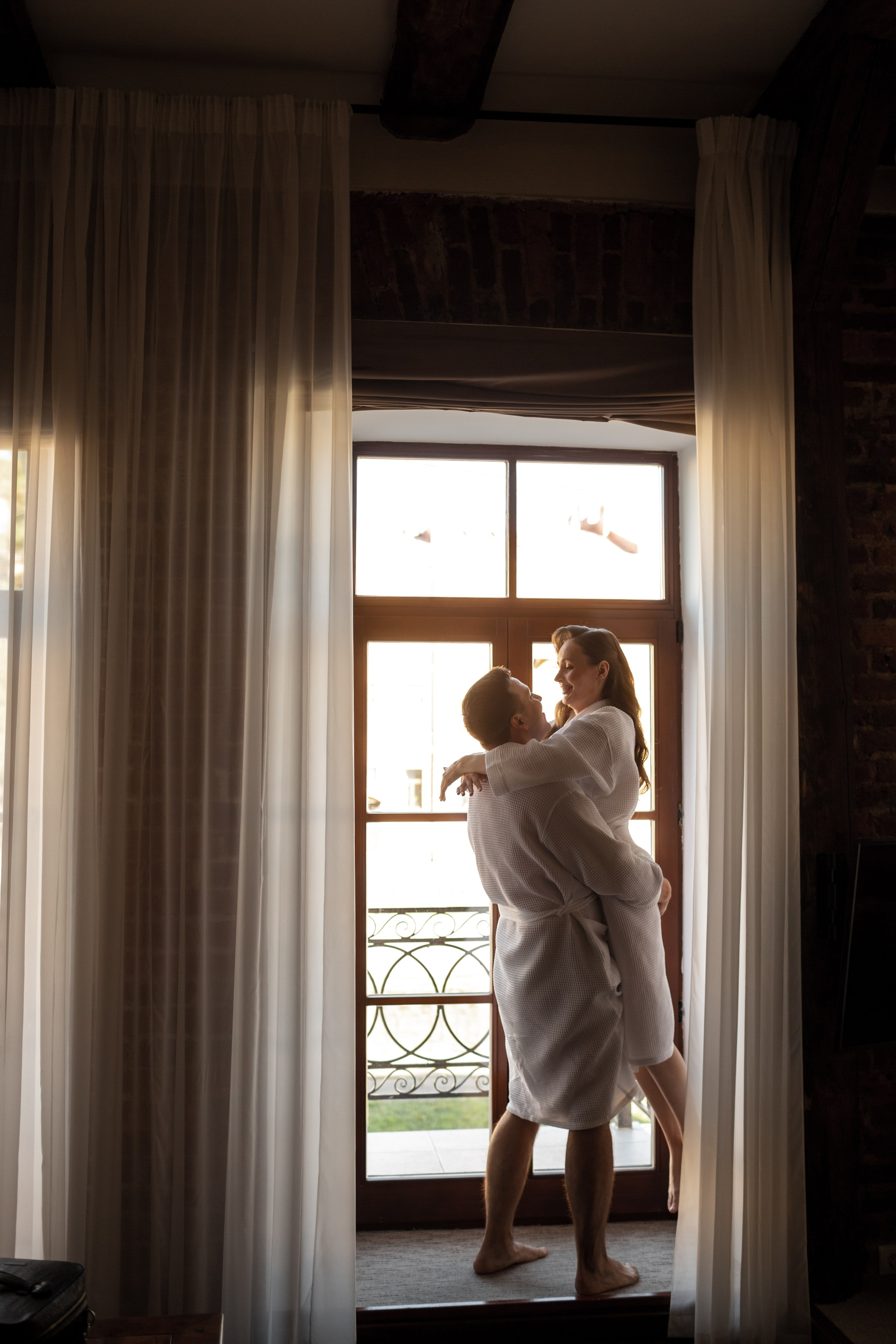 Свадебное утро в отеле: жених обнимает невесту у панорамного окна.