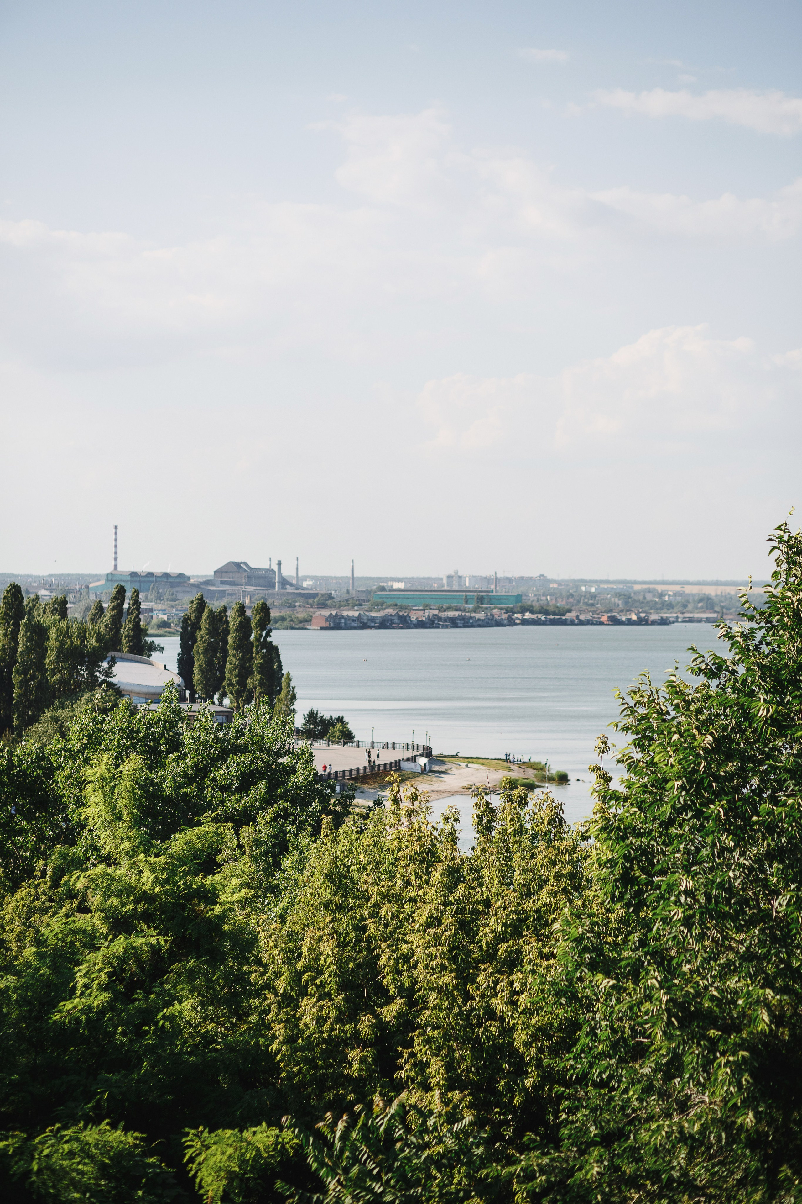 Вид на Пушкинскую набережную в Таганроге с зелеными деревьями на переднем плане и промышленными зданиями вдали. На заднем плане виднеется Таганрогский залив и городской пейзаж.