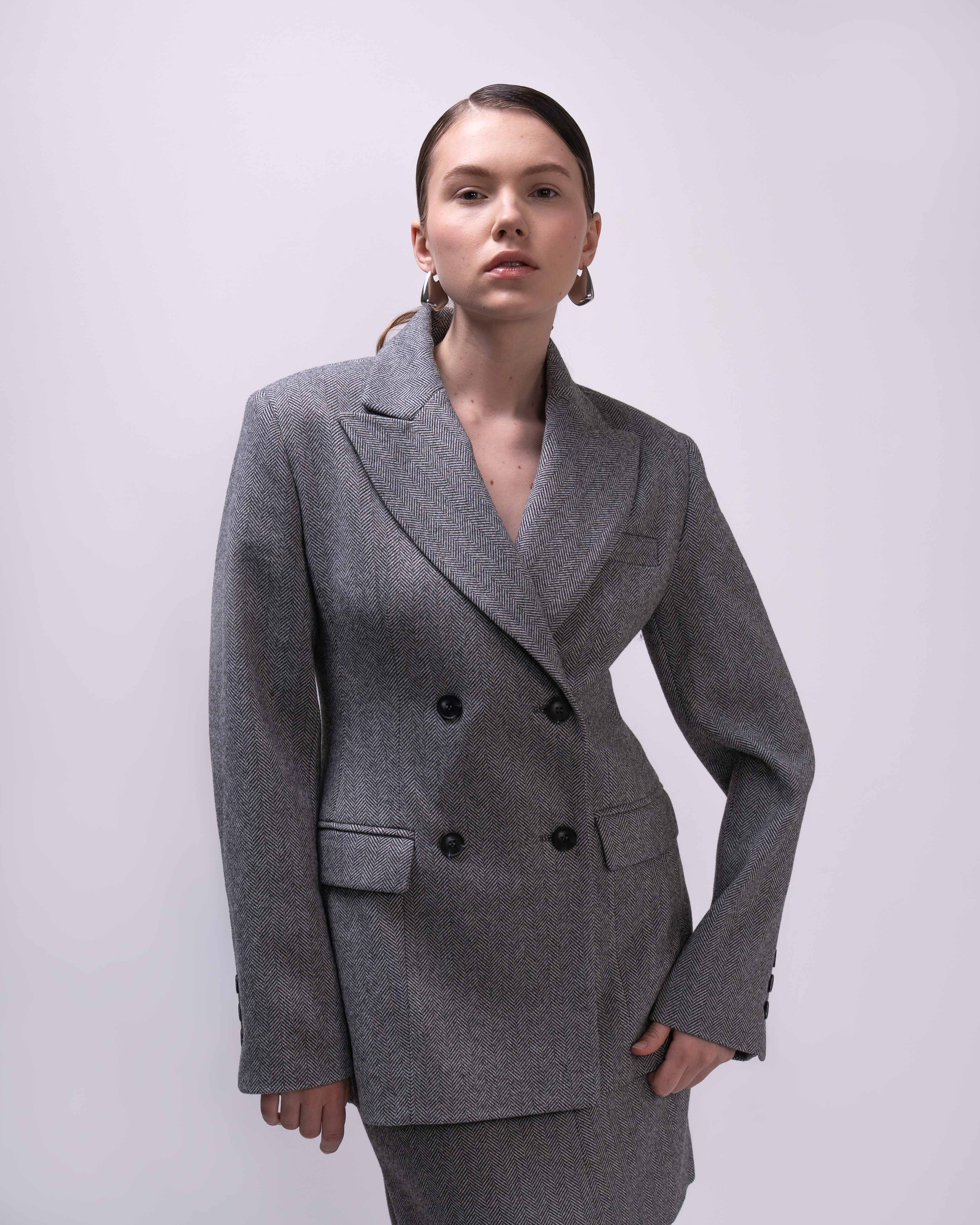 Фото товара для каталога  Ozon или Wildberries.  На фотографии изображена молодая женщина, стоящая на фоне однотонной стены. Она одета в серый пиджак с узором в клетку, который застегнут на две черные пуговицы. Пиджак имеет классический крой с лацканами и карманами. Женщина смотрит в сторону камеры, её взгляд спокойный и уверенный. Волосы у неё собраны в аккуратный пучок, а на ушах видны серьги. Фон изображения нейтральный, что позволяет сосредоточить внимание на модели и её наряде.