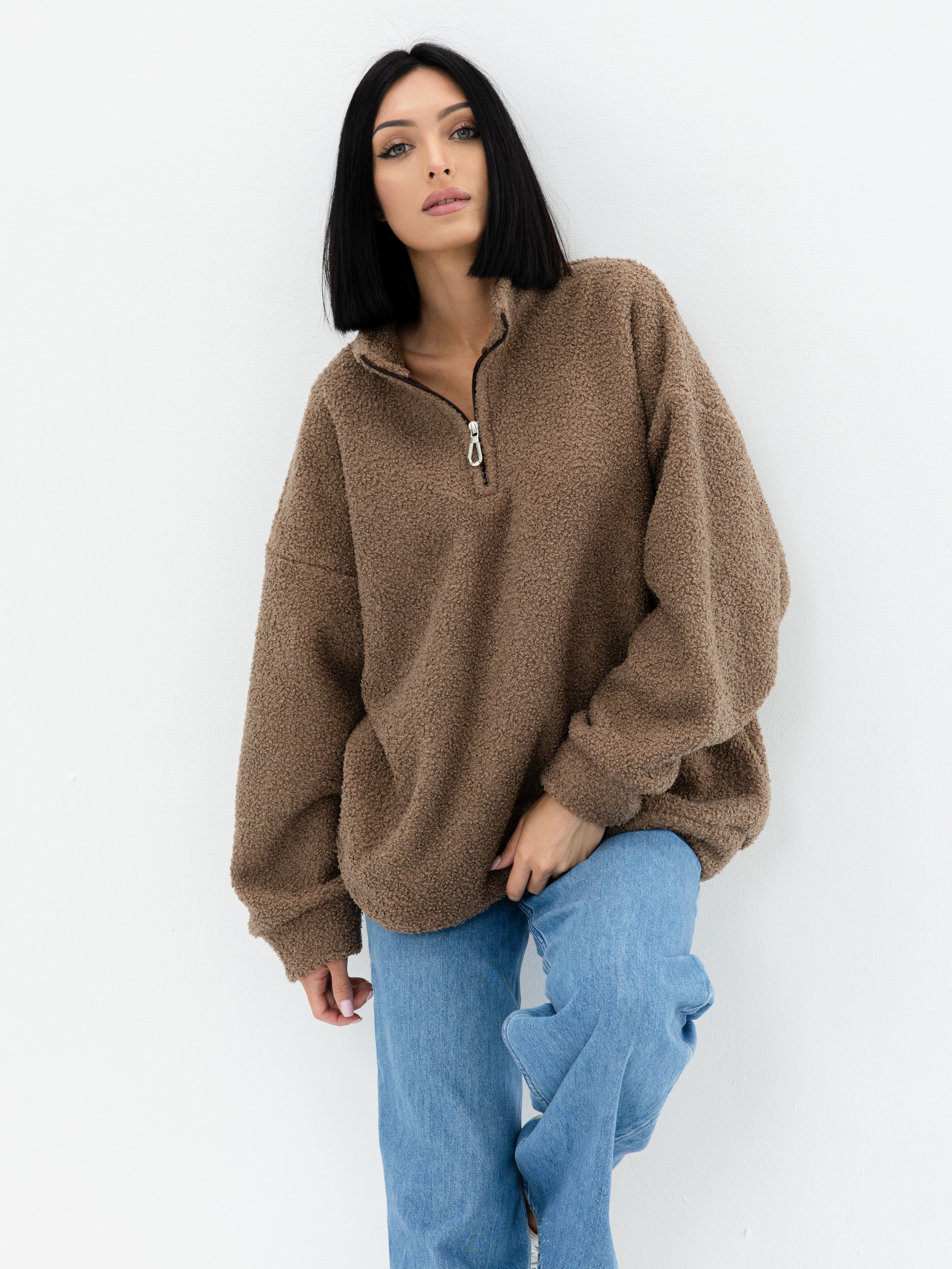 Девушка брюнетка с зелеными глазами в бежевом свитере букле на молнии и джинсах стоит на белом фоне