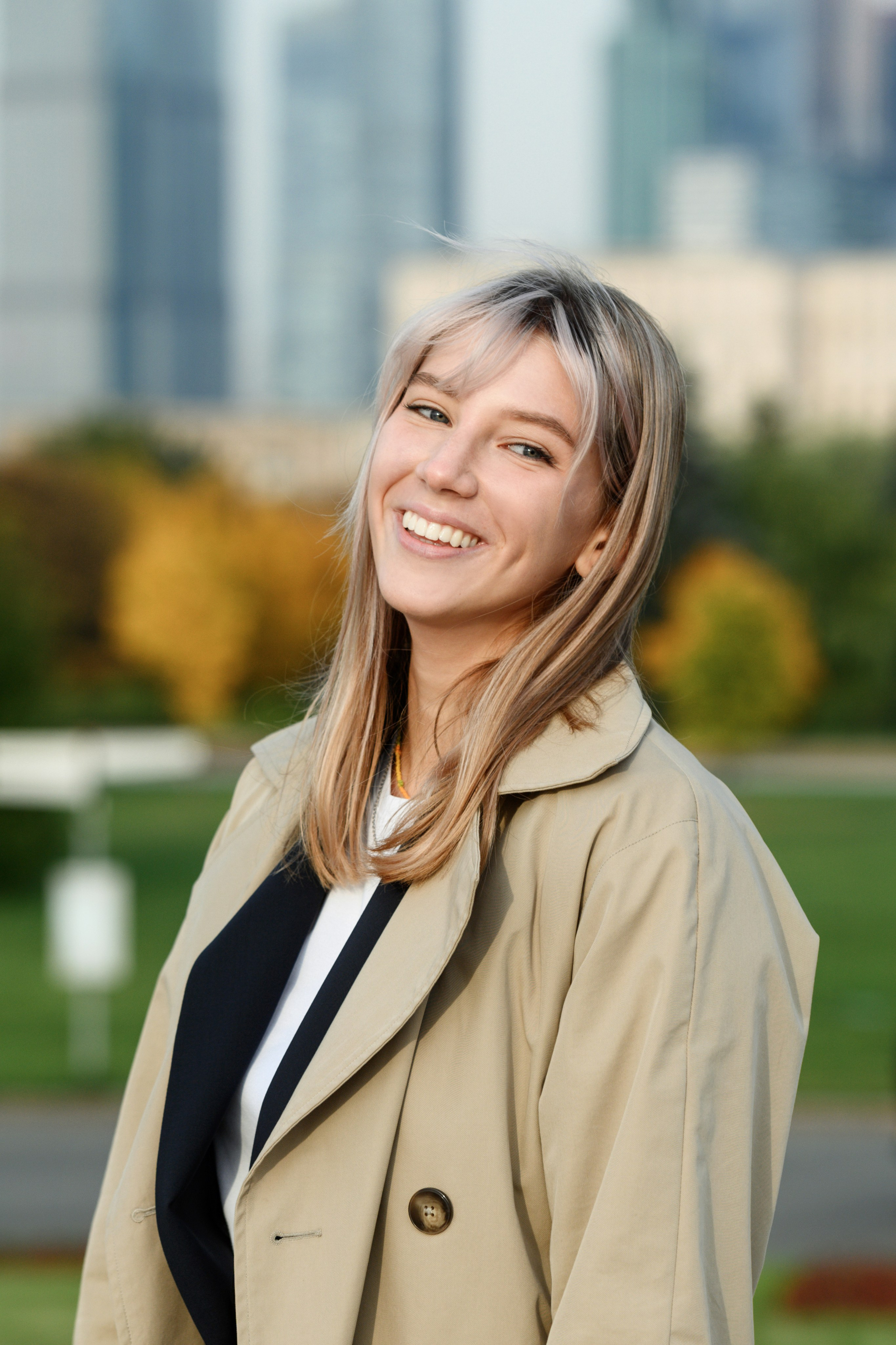 Улыбающийся уличный портрет выпускницы в бежевом тренче на фоне осеннего парка и небоскрёбов.