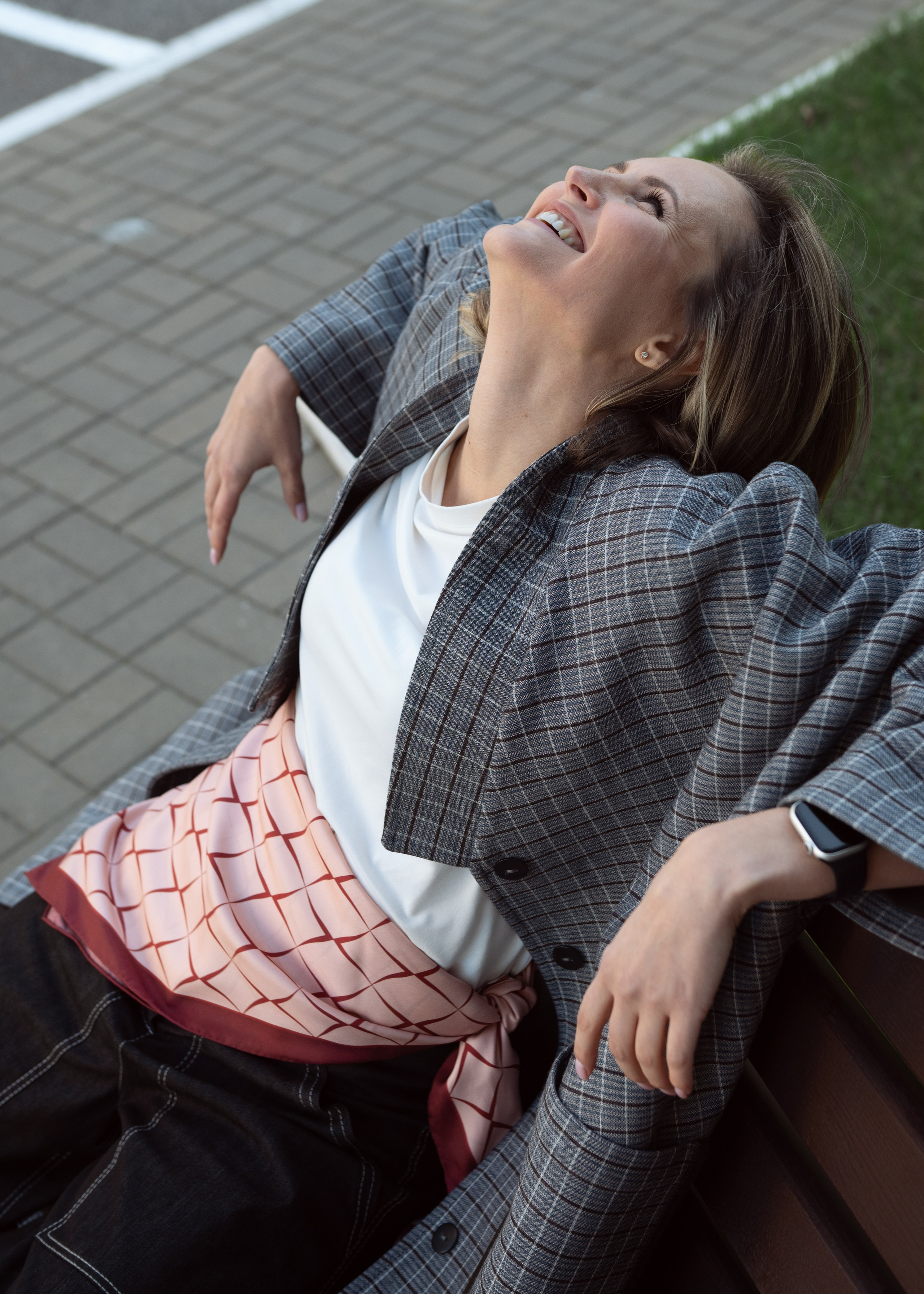 Эмоциональный женский портрет: Женщина сидит на скамейке запрокинув голову и улыбается. На ней белая футболка, серое пальто в клетку, черные брюки и роскошный розовый платок на поясе. Съемка на улице для контента магазина женской одежды и аксессуаров.