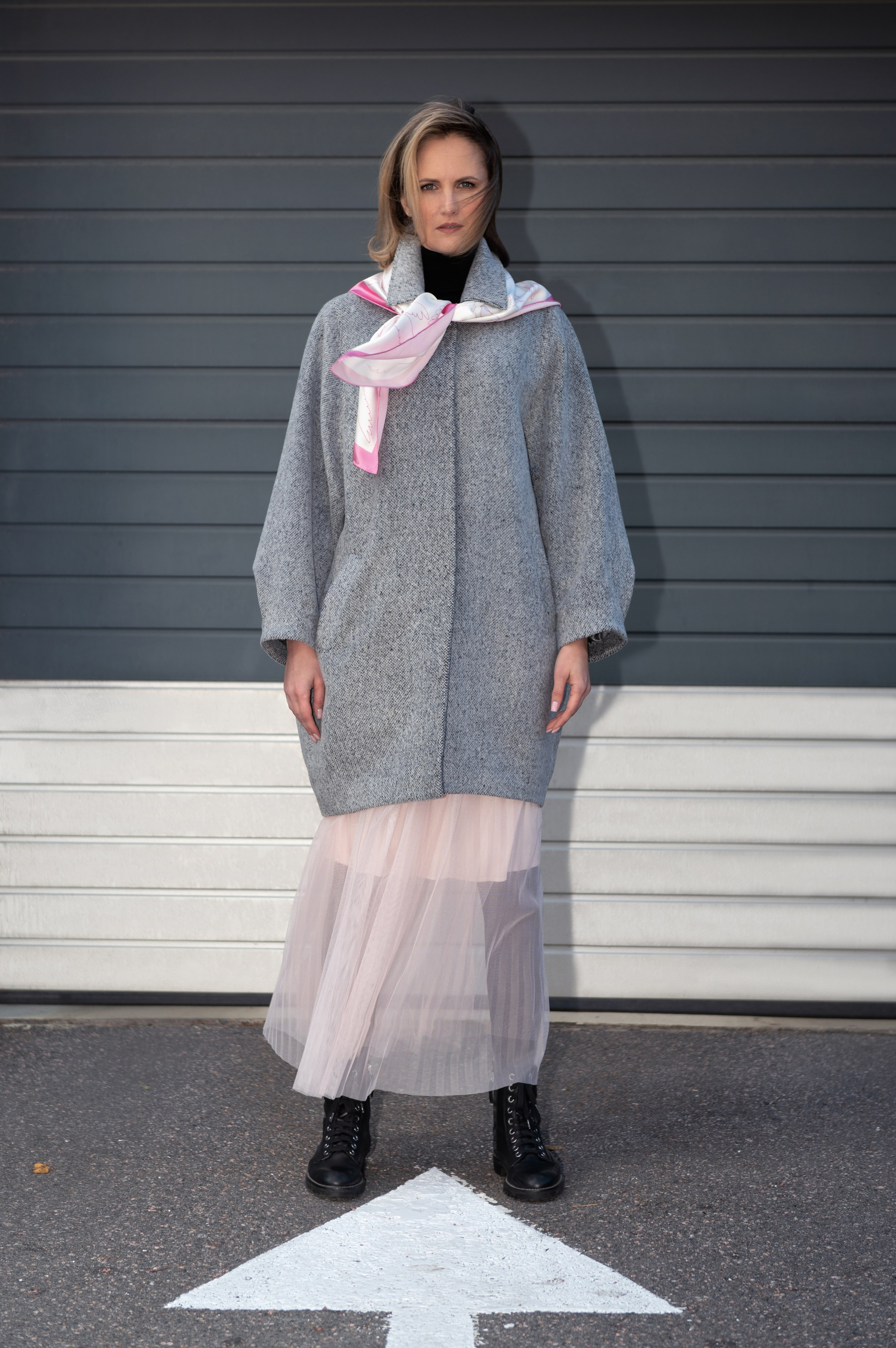 Женщина в сером пальто-коконе,черной водолазке, розовой юбке из фатина и грубых черных ботинках стоит у серо-белой стены паркинга, на шее у нее бело-розовый шелковый платок. Съемка со смешанным светом. Использован прием "вспышка в лоб", чтобы выделить модель из фона.