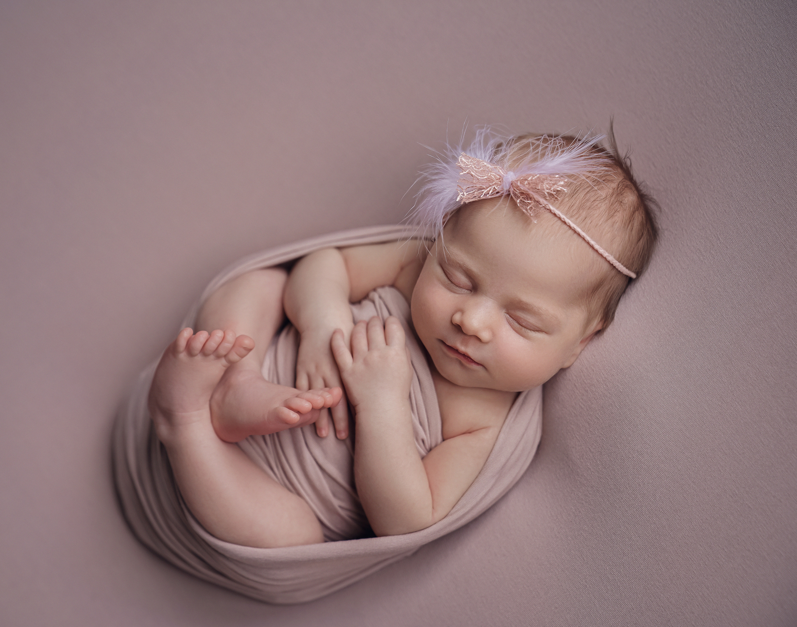 новорожденная девочка на фотографии уютно лежит, свернувшись как калачик с красивой повязкой на голове