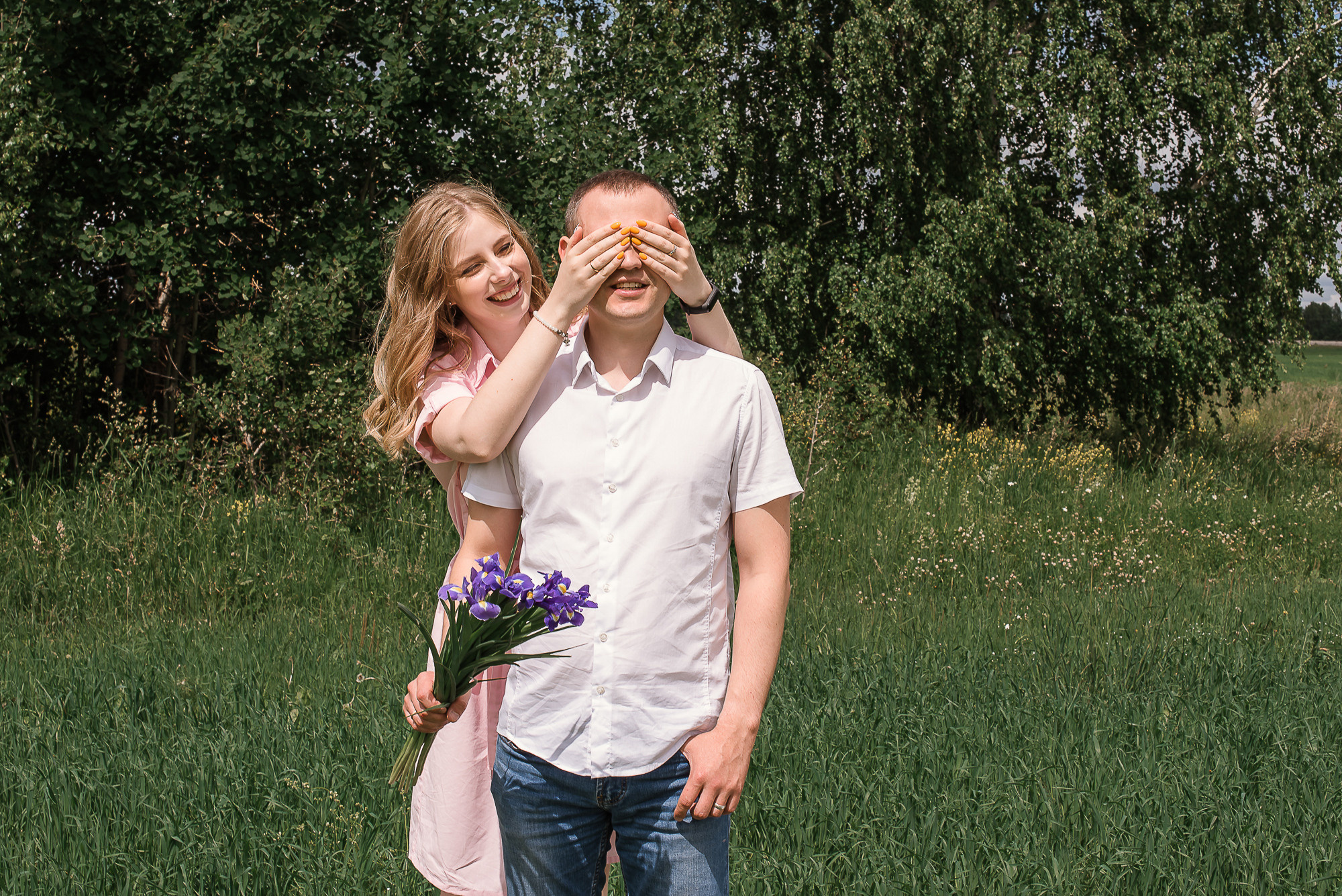 девушка в розовом платье закрыла глаза, парень в рубашке стоят на фоне зеленых деревьев с цветами, фотосессия в поле, фотосессия летом