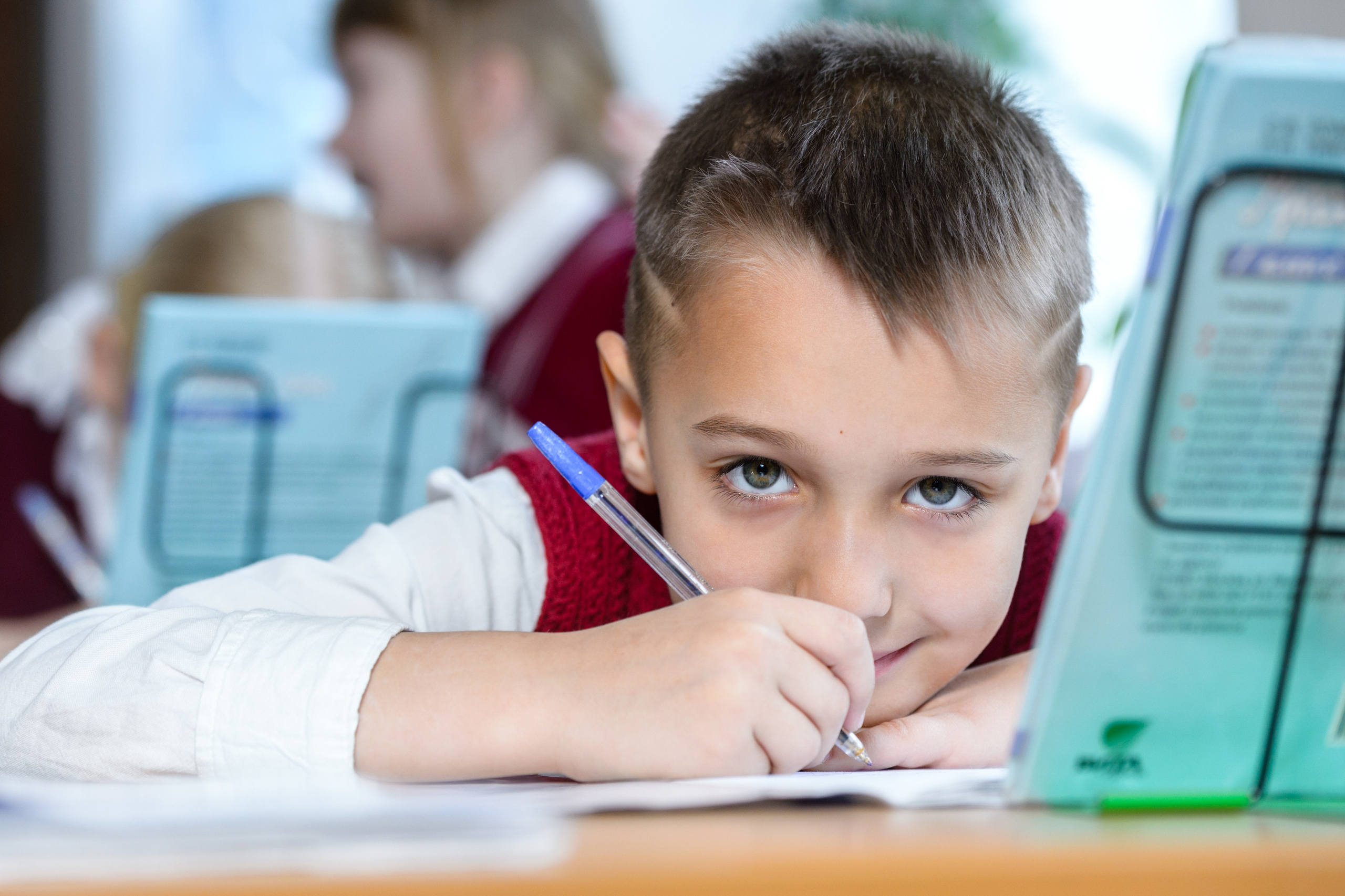 Мальчик за партой в классе смотрит в камеру, опираясь на руку рядом с учебными принадлежностями.
