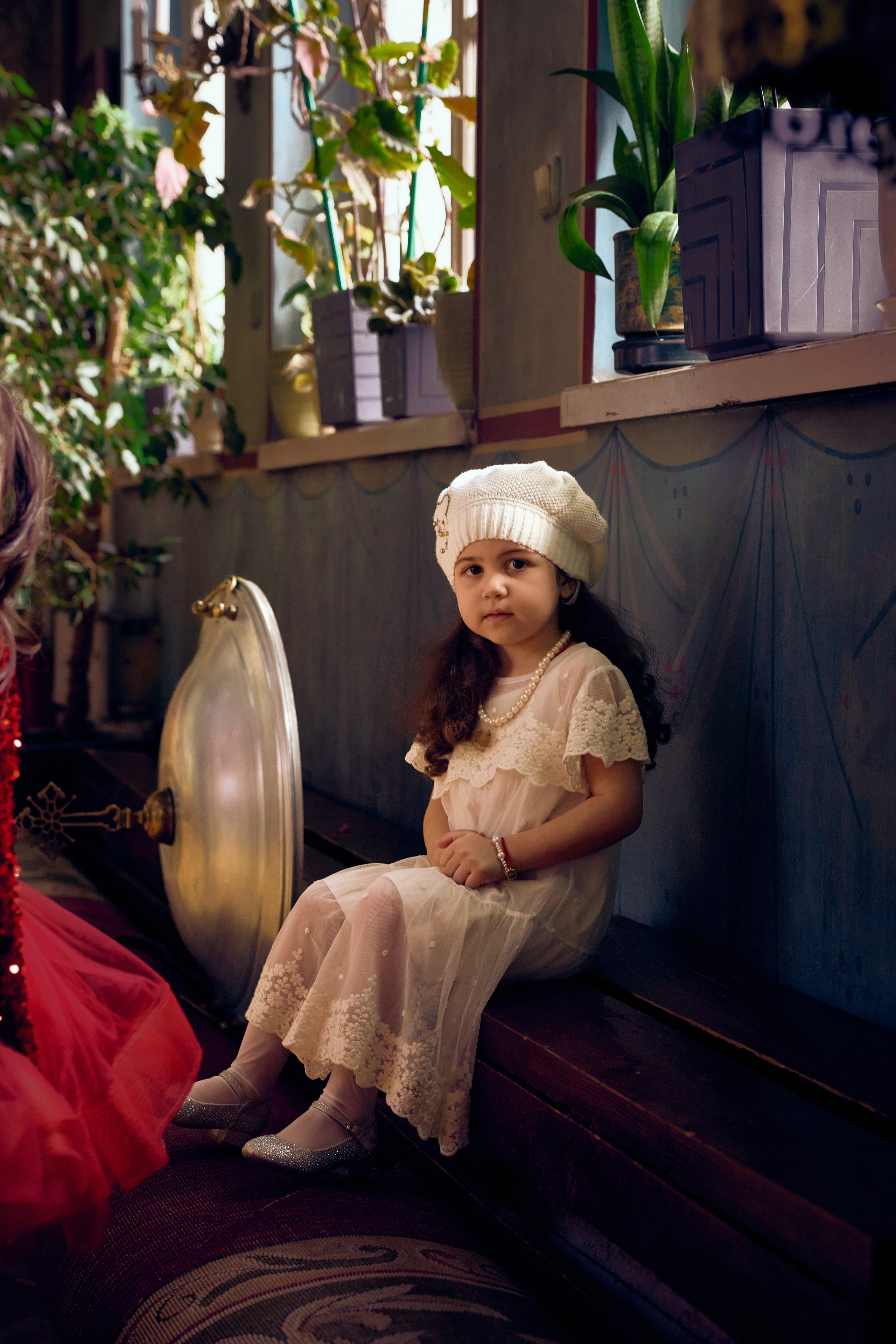 Маленькая девочка в белом платье и шапочке сидит на скамейке у окна в православной церкви