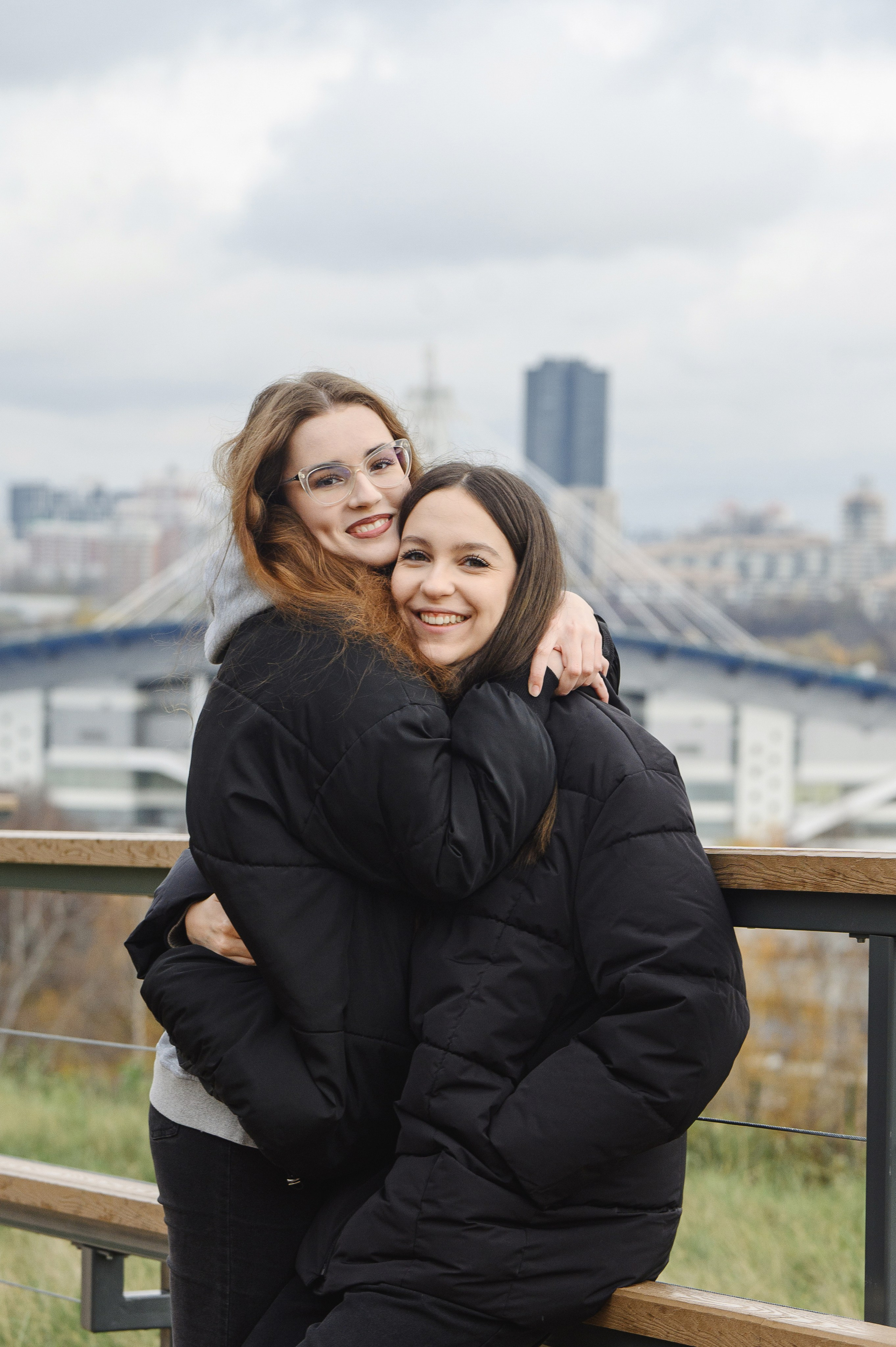 Две подруги в тёмной верхней одежде обнимаются на фоне города.