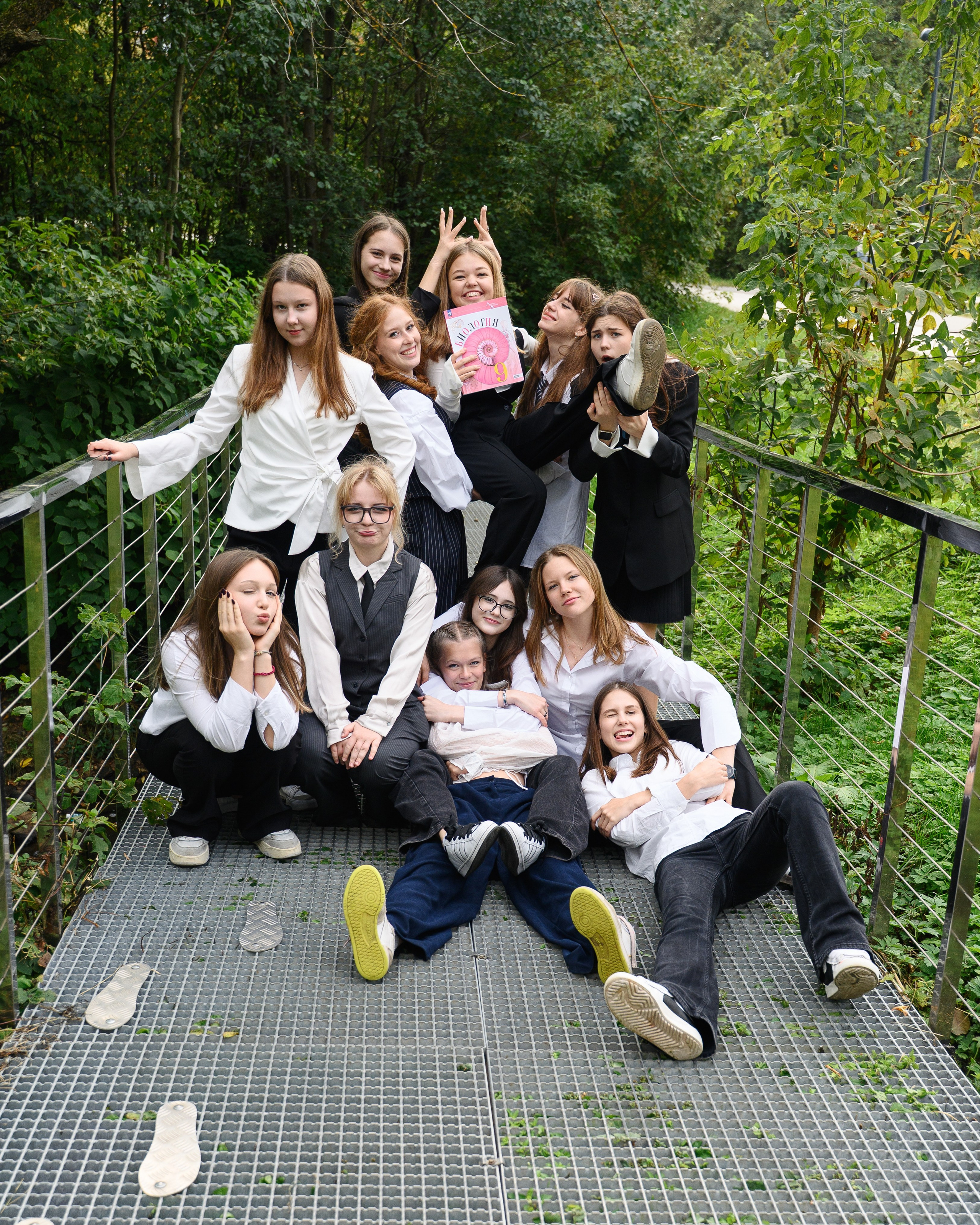 Компания одноклассниц сидит и стоит на мостике в парке в живом неформальном кадре.