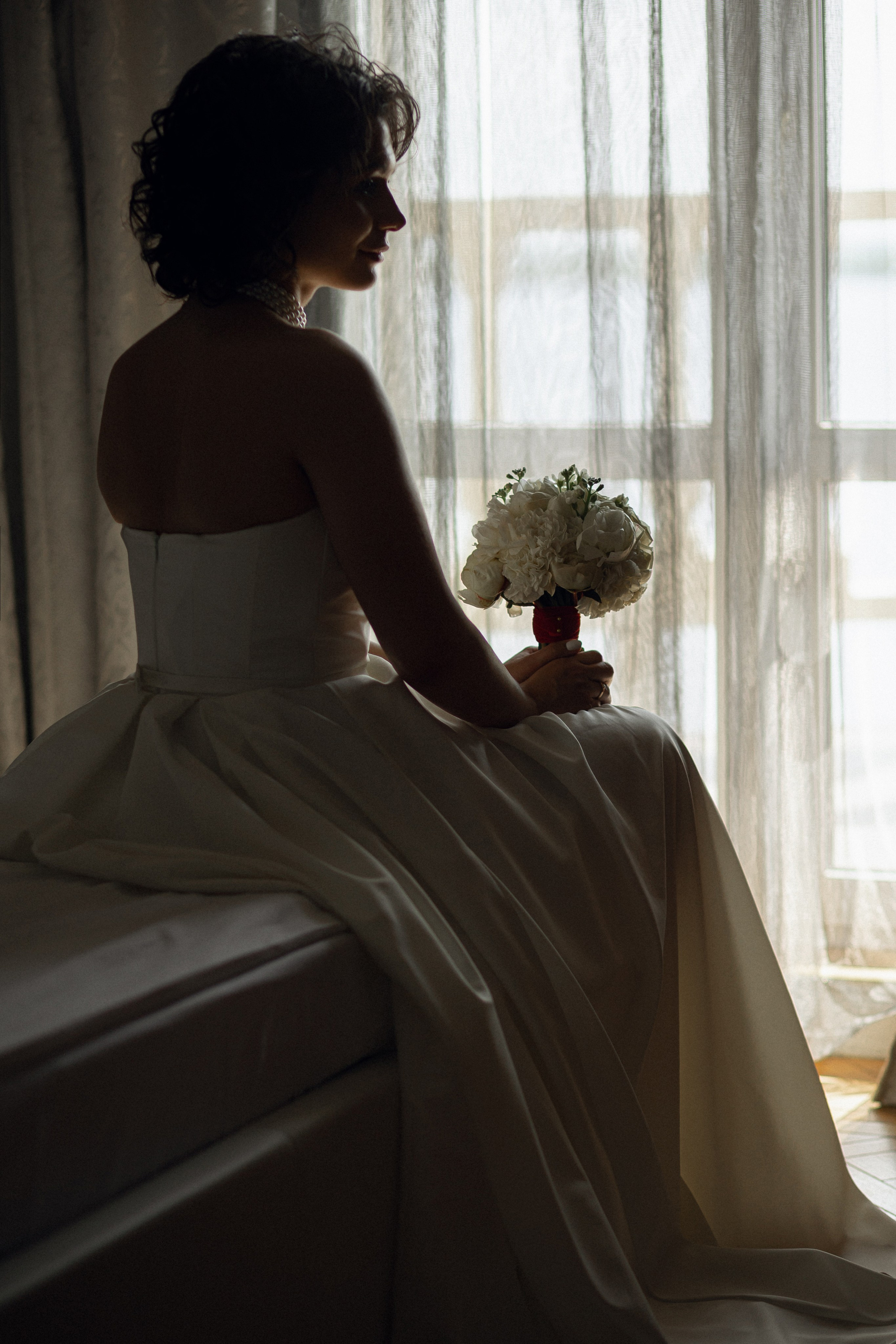 Романтический кадр для портфолио: невеста в атласном платье задумчиво смотрит на букет роз в полумраке спальни с деревянной кроватью и шелковым постельным бельем