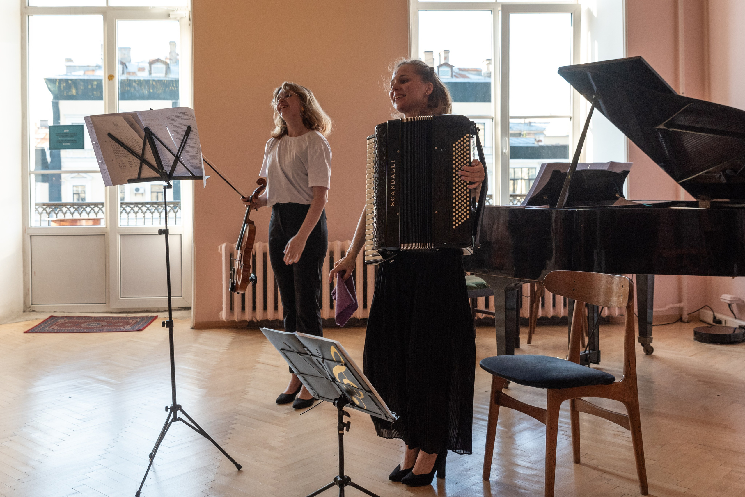 Девушки музыканты закончили выступление, в руках скрипка и аккордеон, за спиной рояль, яркий солнечный свет из окон.