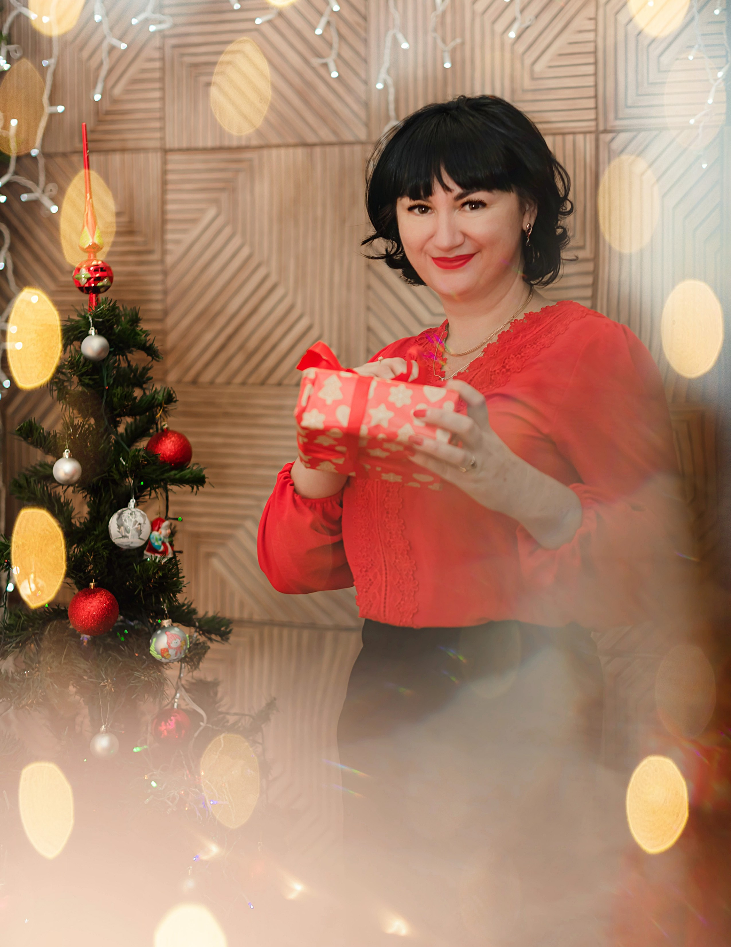 На фото женщина с подарком на новый год в студии в красной блузке около елочки.