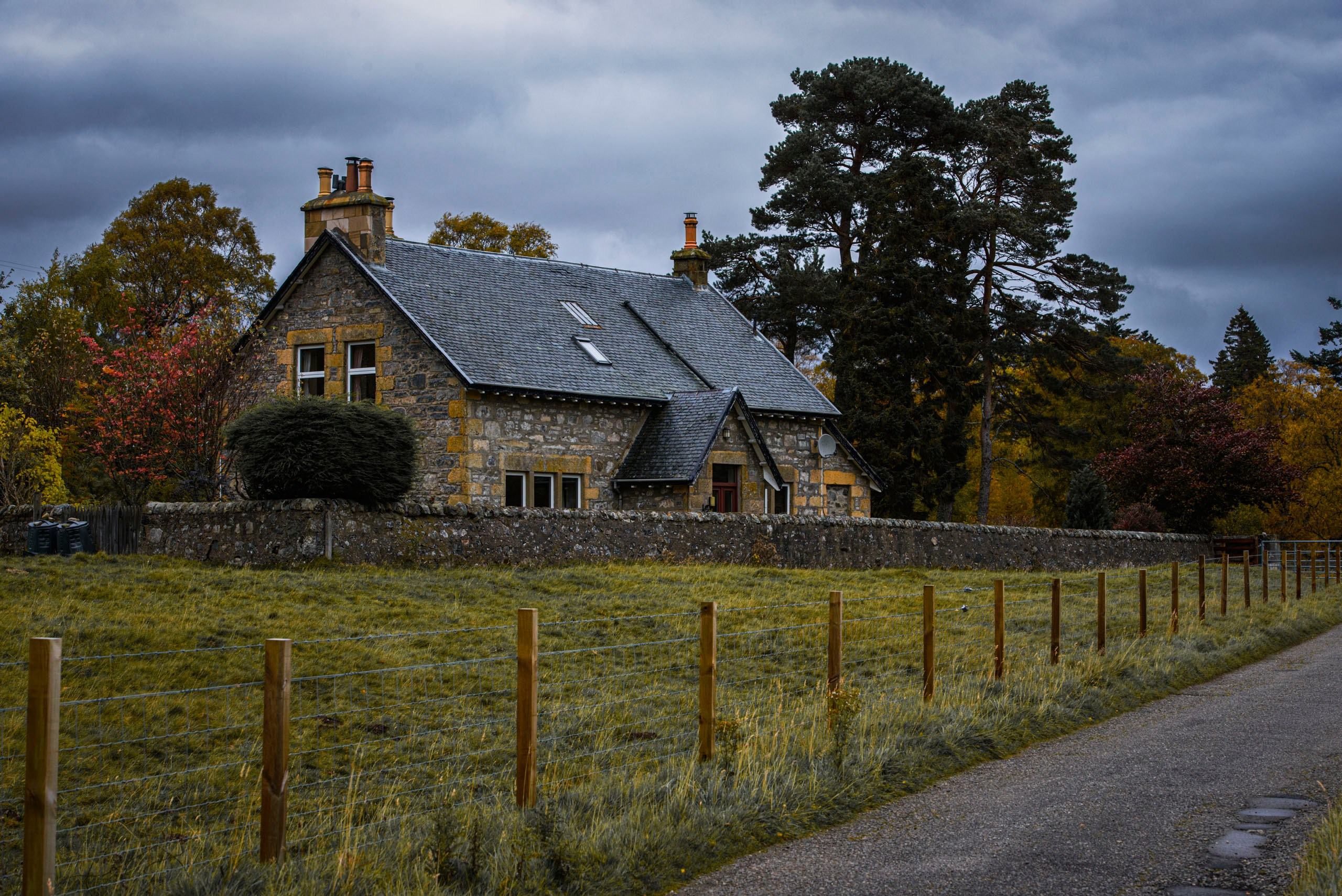 Традиционный шотландский каменный коттедж с серой крышей и каменной стеной, окруженный осенними деревьями под пасмурным небом.