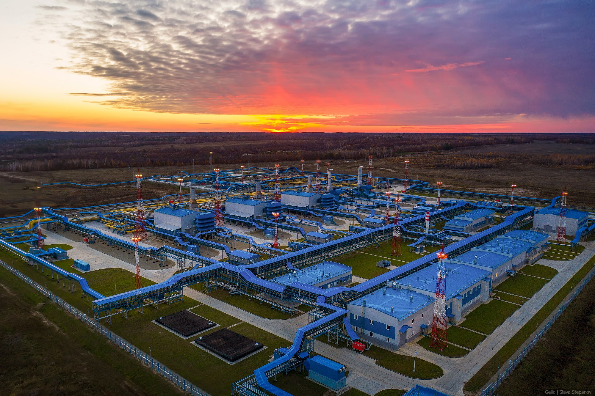 Газокомпрессорная станция магистрального газопровода на ярком закате, гигантская сеть синих трубопроводов и цехов с высоты птичьего полёта, Gelio.
