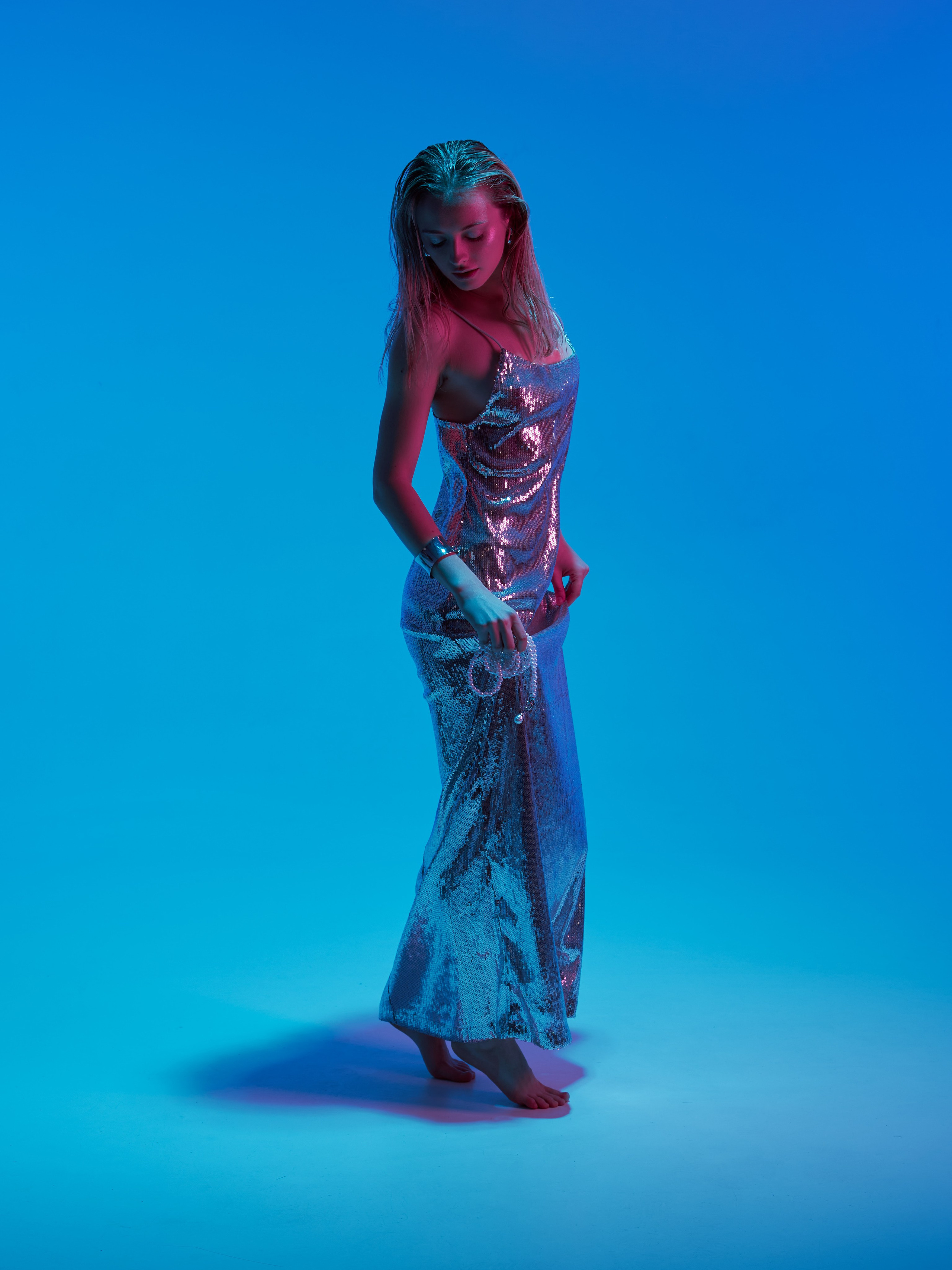 Девушка в длинном серебристом платье на тонких бретельках в синем свете на белой циклопаме
