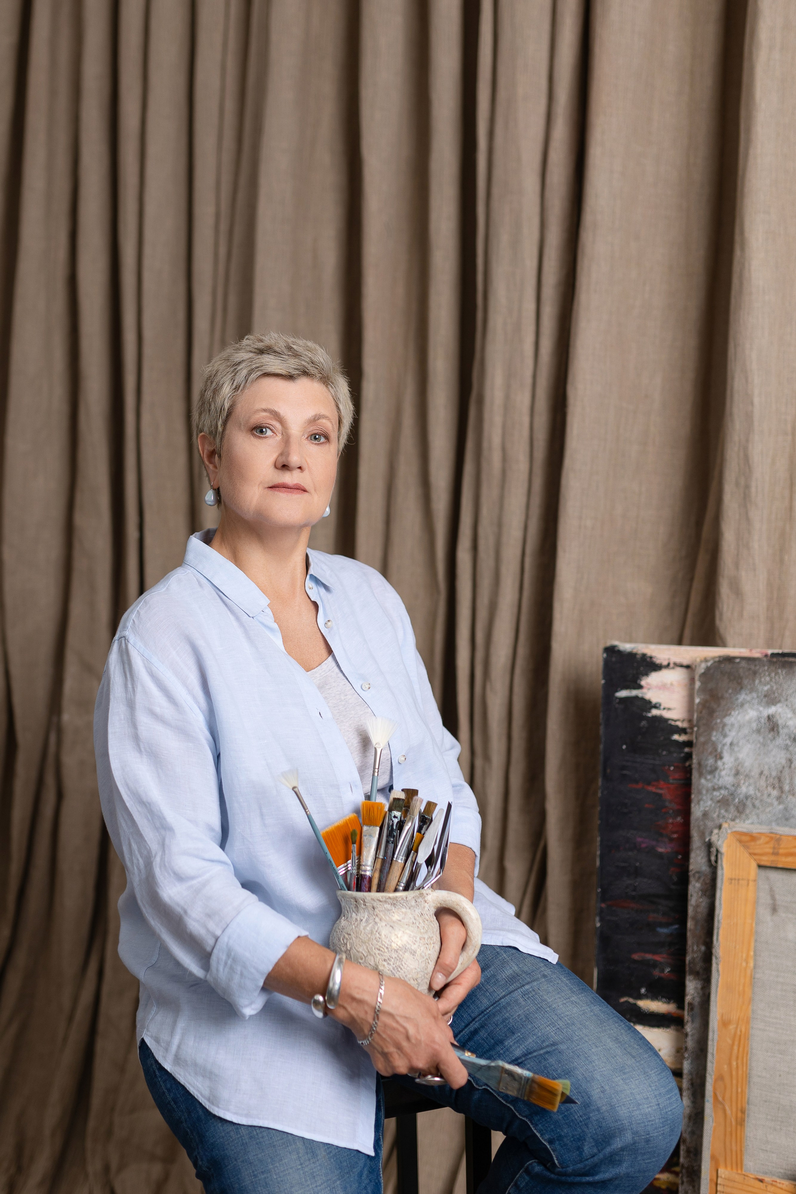 Художница сидит  на фоне серых штор, держа в  руках вазу с кистями.