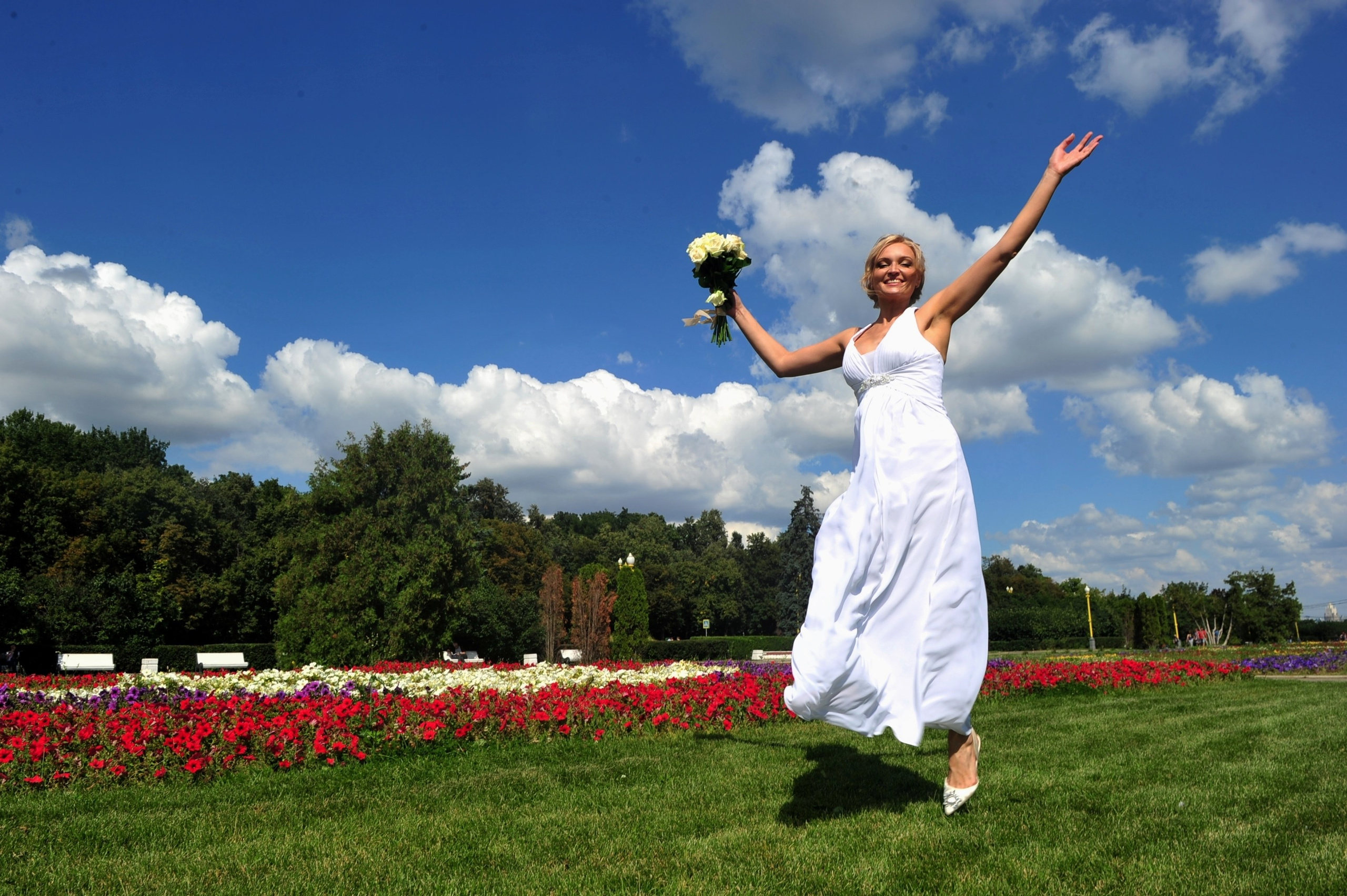 Свадьба на Воробьёвых Горах рядом с МГУ, невеста со свадебным букетом из кремовых роз радостно прыгает на зеленой лужайке на фоне синего неба, красных и белых цветов