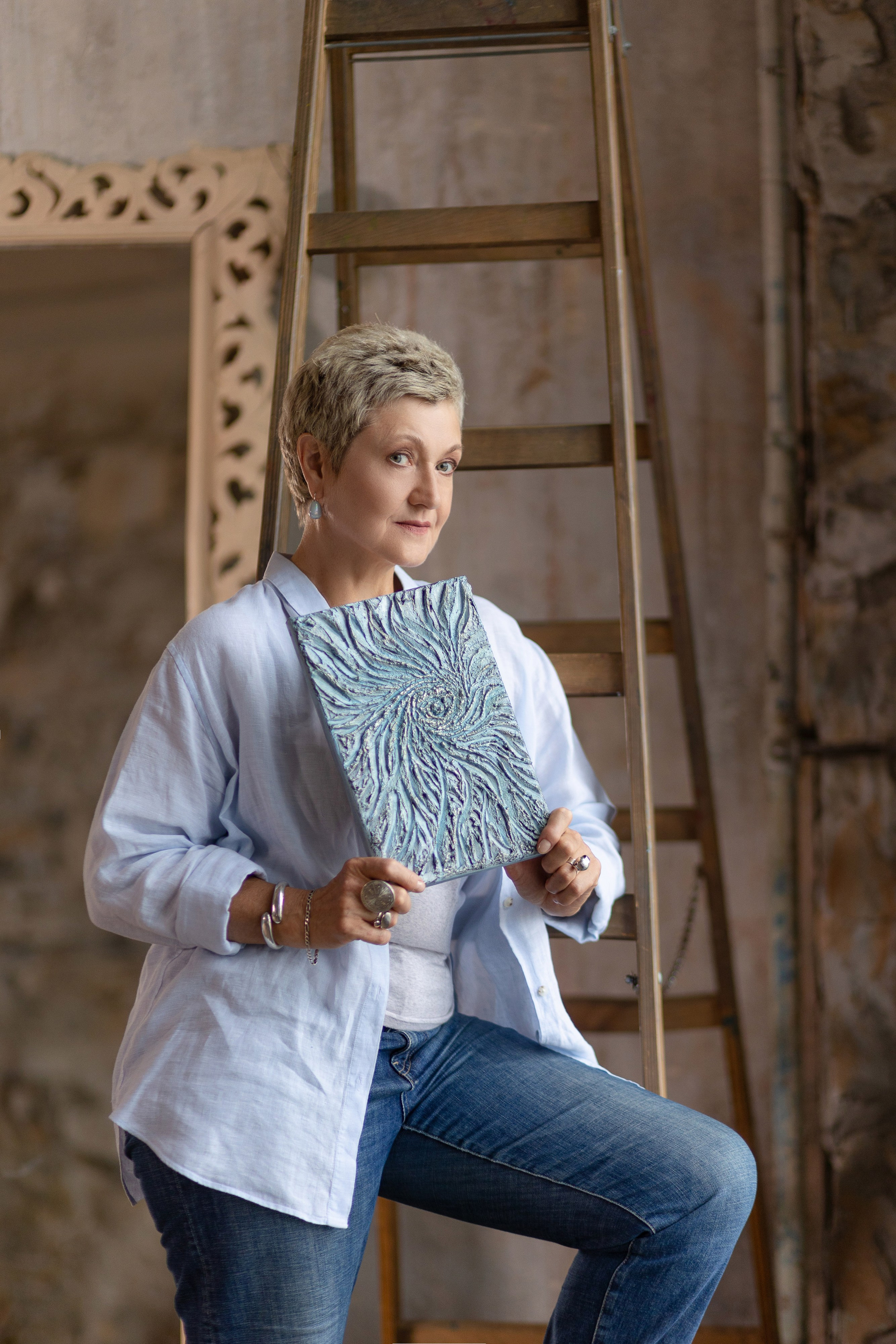 Художница стоит на фоне лестницы и большого холста в рамке, держа в руках картину. 