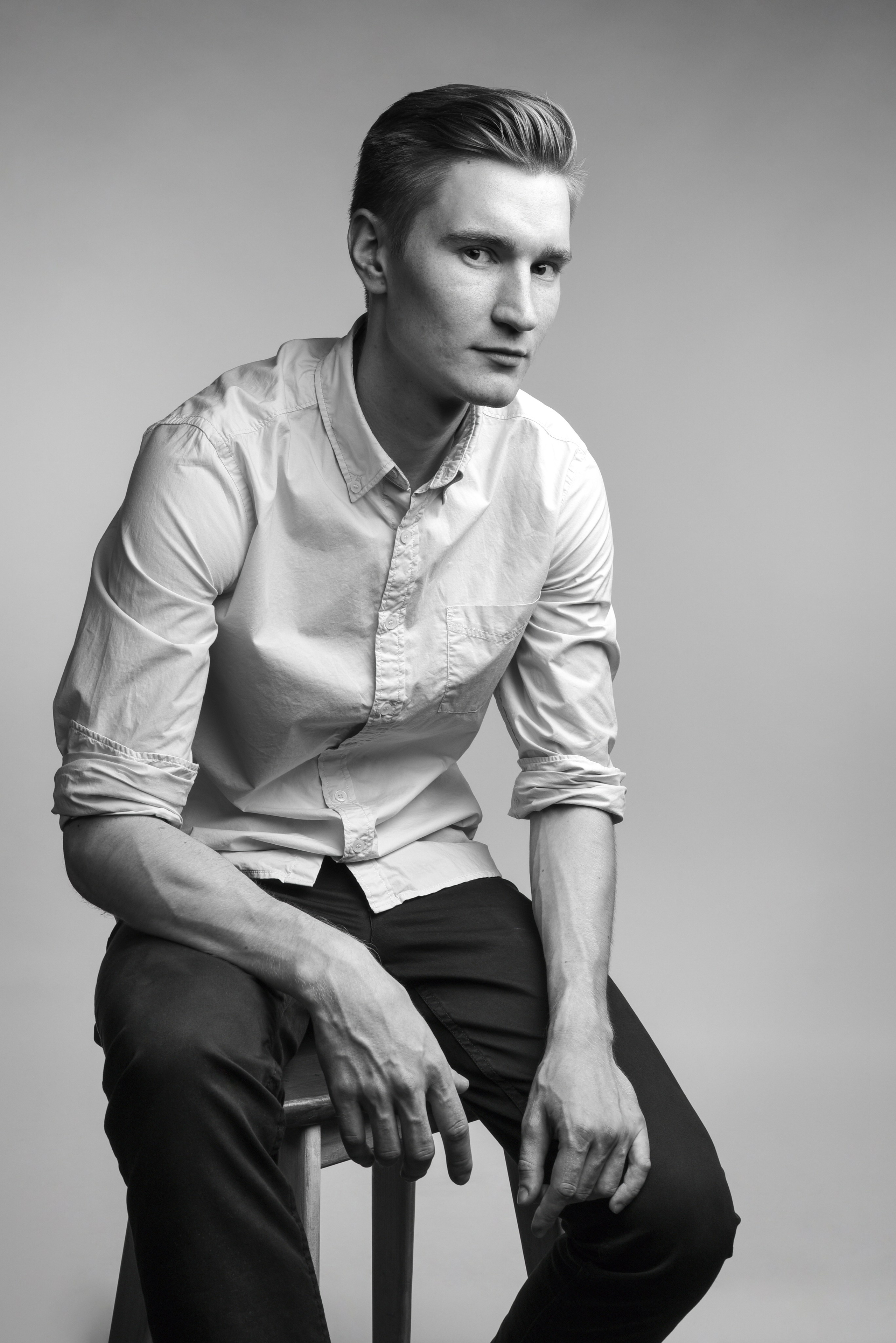 Фотостудия. Молодой человек в белой рубашке сидит на табуретке. Руки свободно лежат на коленях.