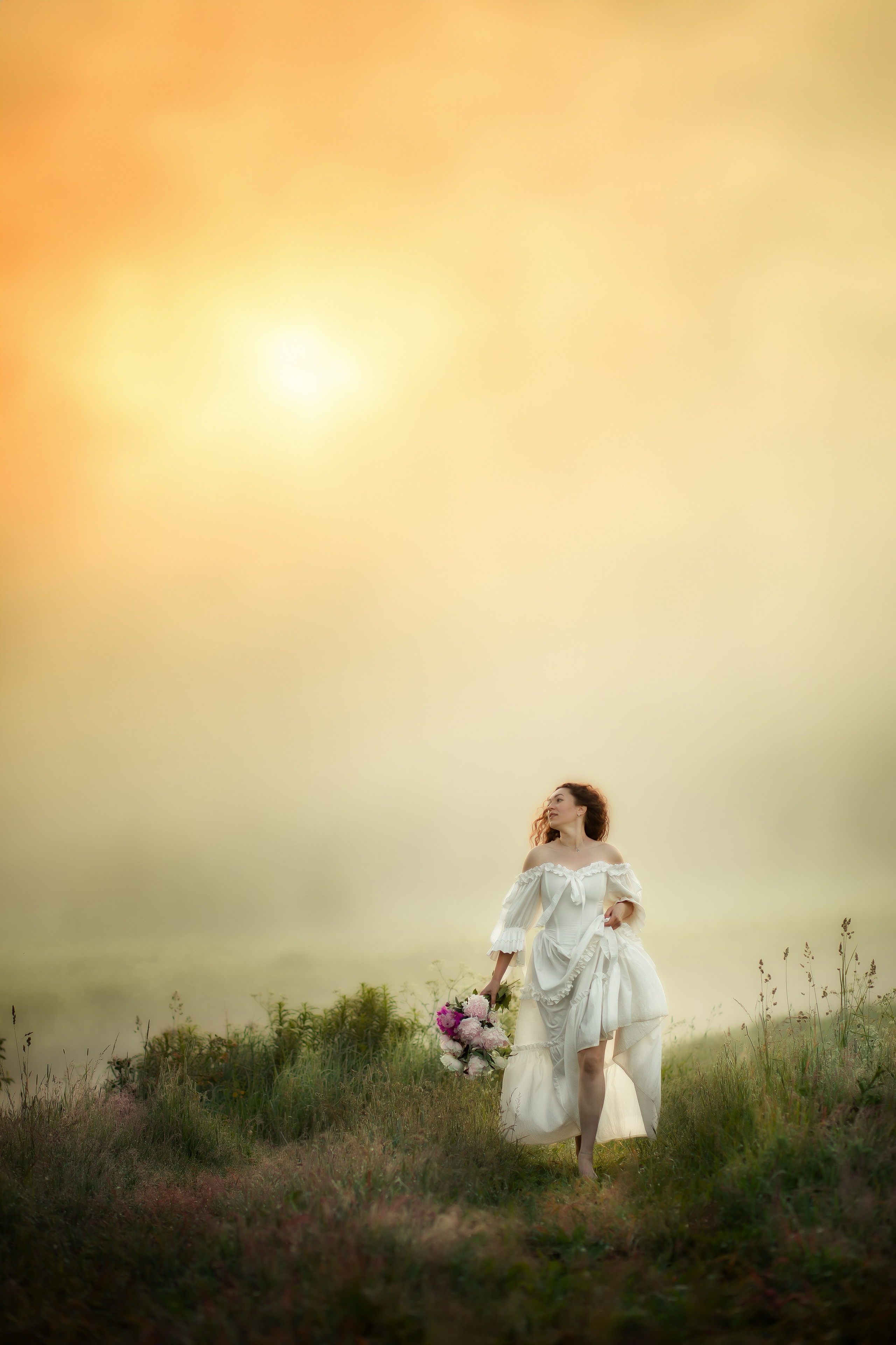 Женщина в белом на фоне туманного рассвета, с букетом в руках и ветром в волосах — в тишине нового дня.