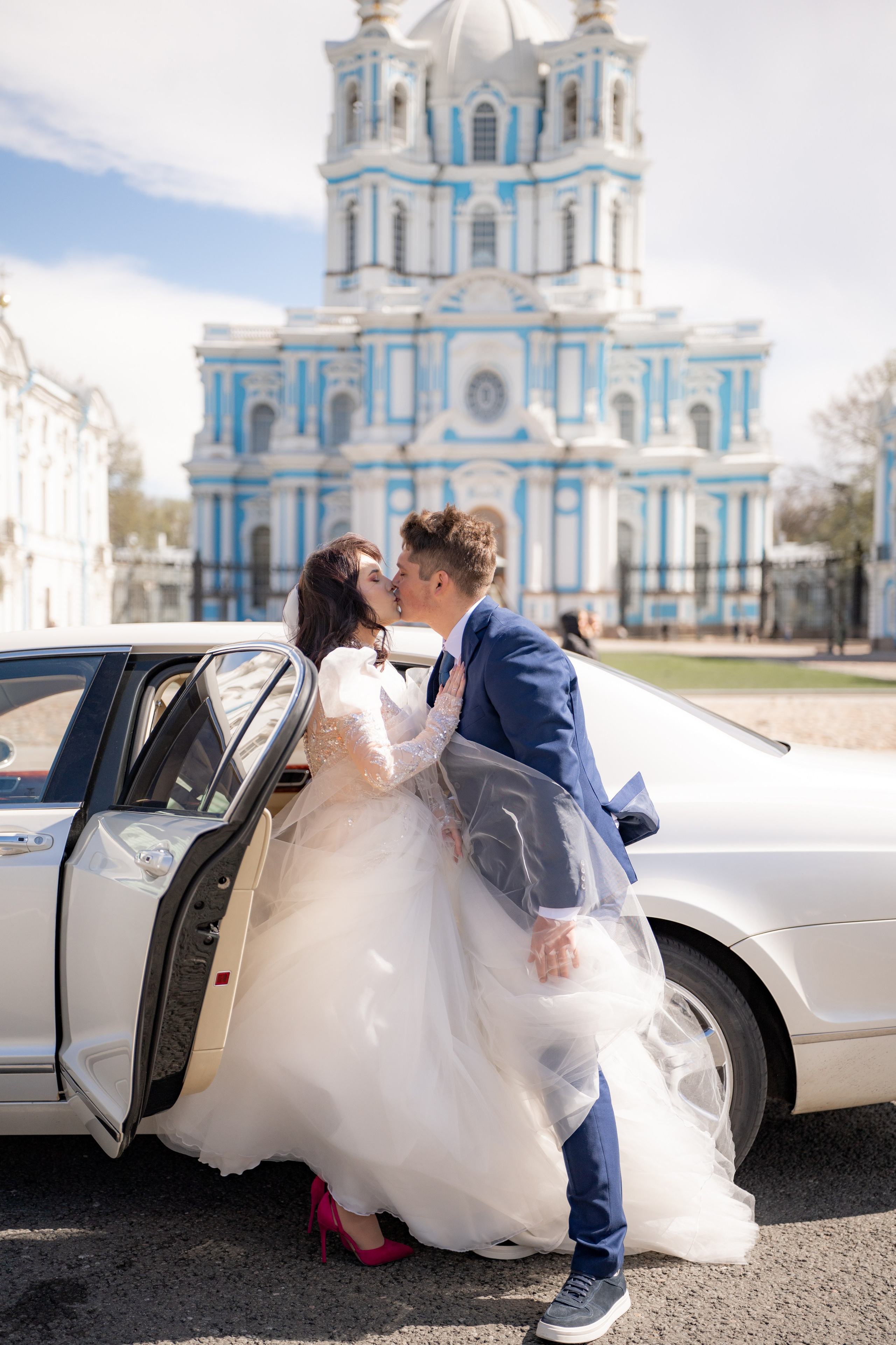 Свадебная фотосессия у Смольного собора. Жених красиво обнимает невесту у автомобиля.