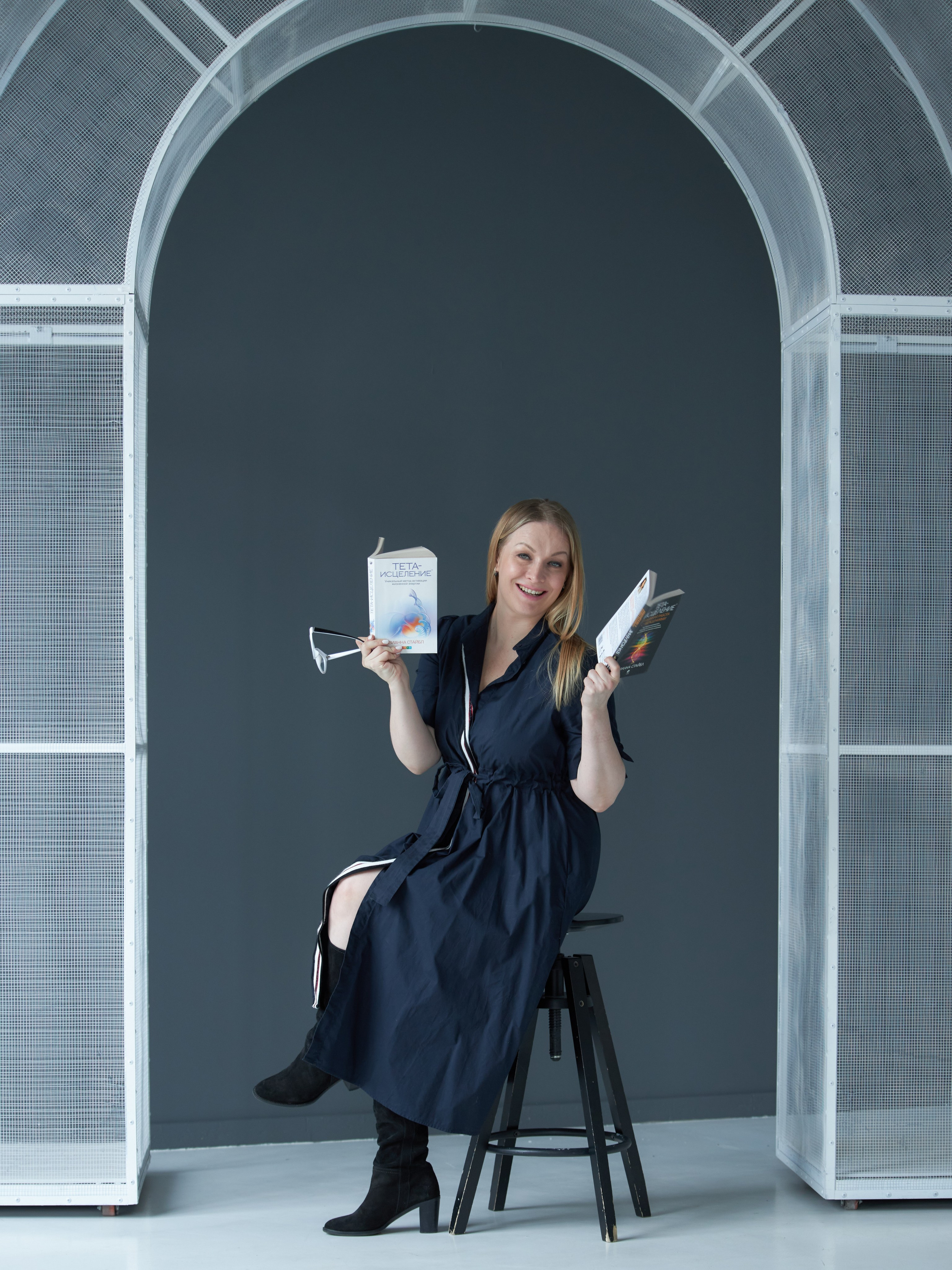 Контент-фотосессия для космоэнергета: девушка в очках и платье сидит на фоне серой стены и держит в руках книги