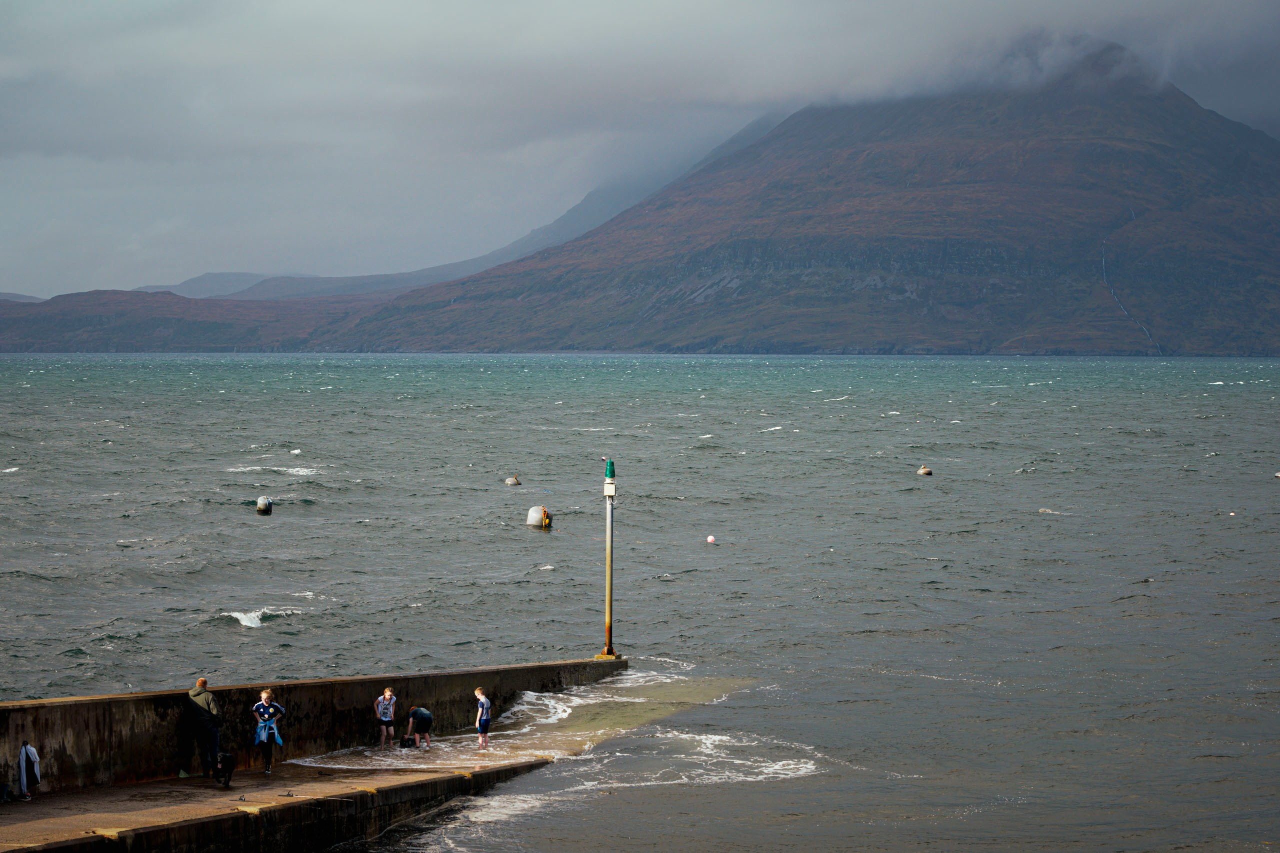 Люди стоят на бетонном пирсе у бурного моря на острове Скай, Шотландия, с большой темной горой на заднем плане, скрытой облаками.