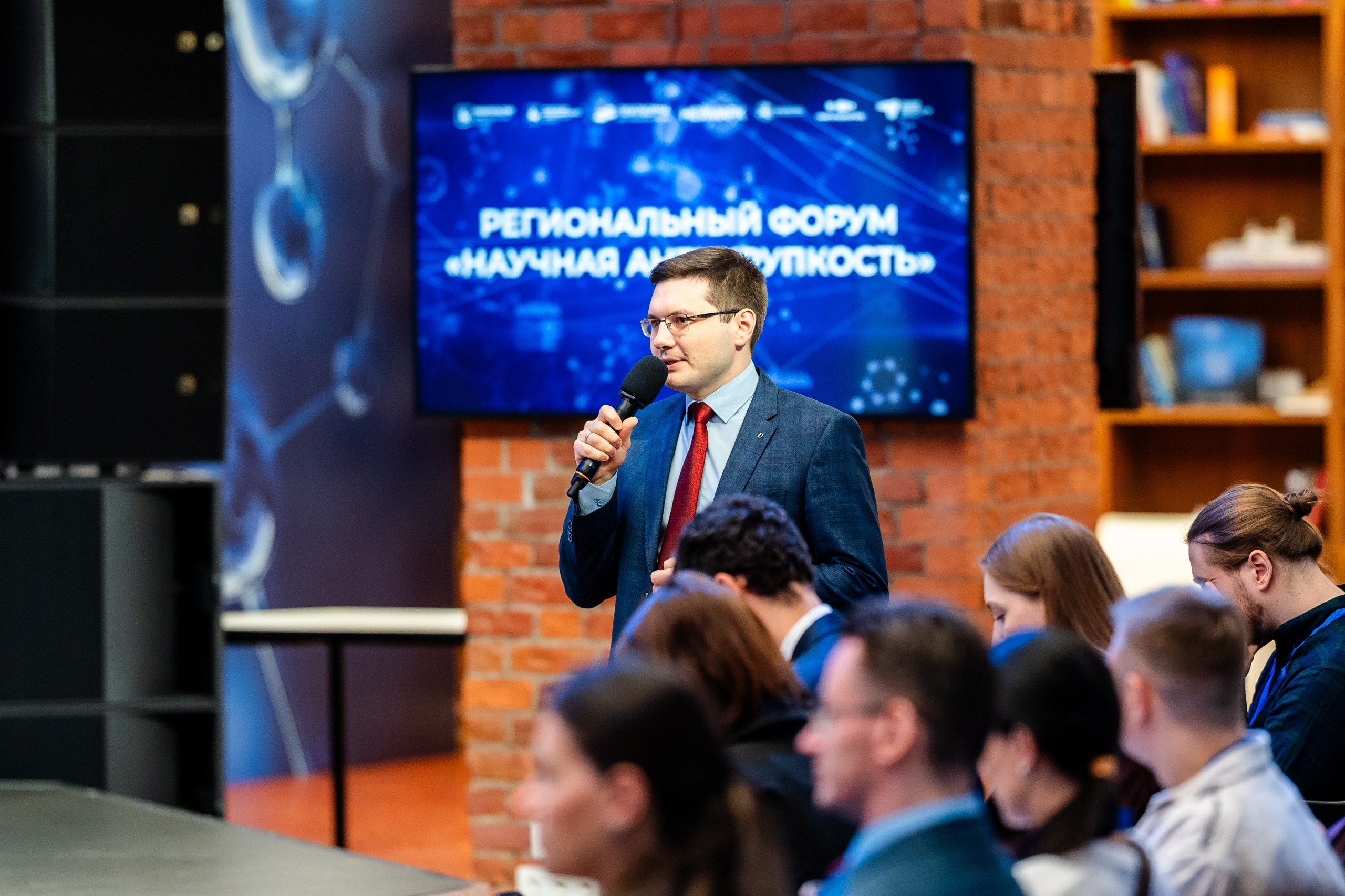 Репортажная съёмка форума: спикер с микрофоном выступает перед аудиторией, экран с надписью «Региональный форум "Научная антихрупкость"»