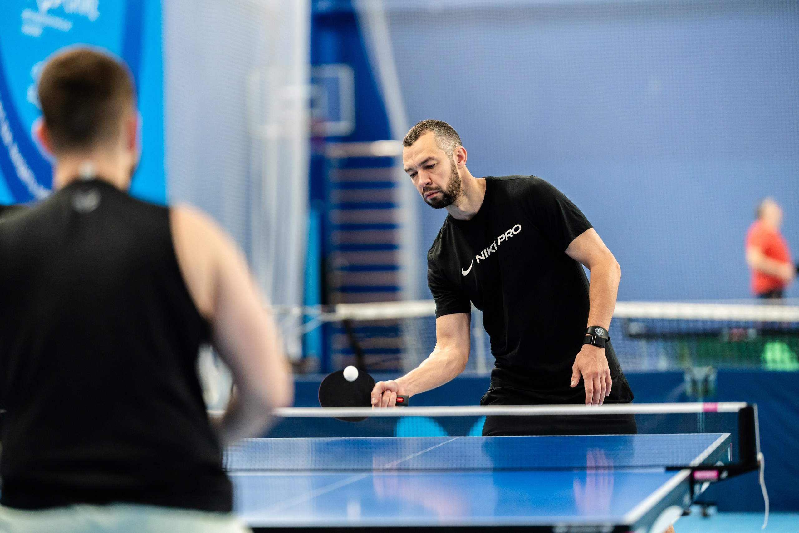 Репортаж с турнира по настольному теннису: спортсмен в футболке Nike Pro с ракеткой и мячом, синий зал и размытый соперник на переднем плане