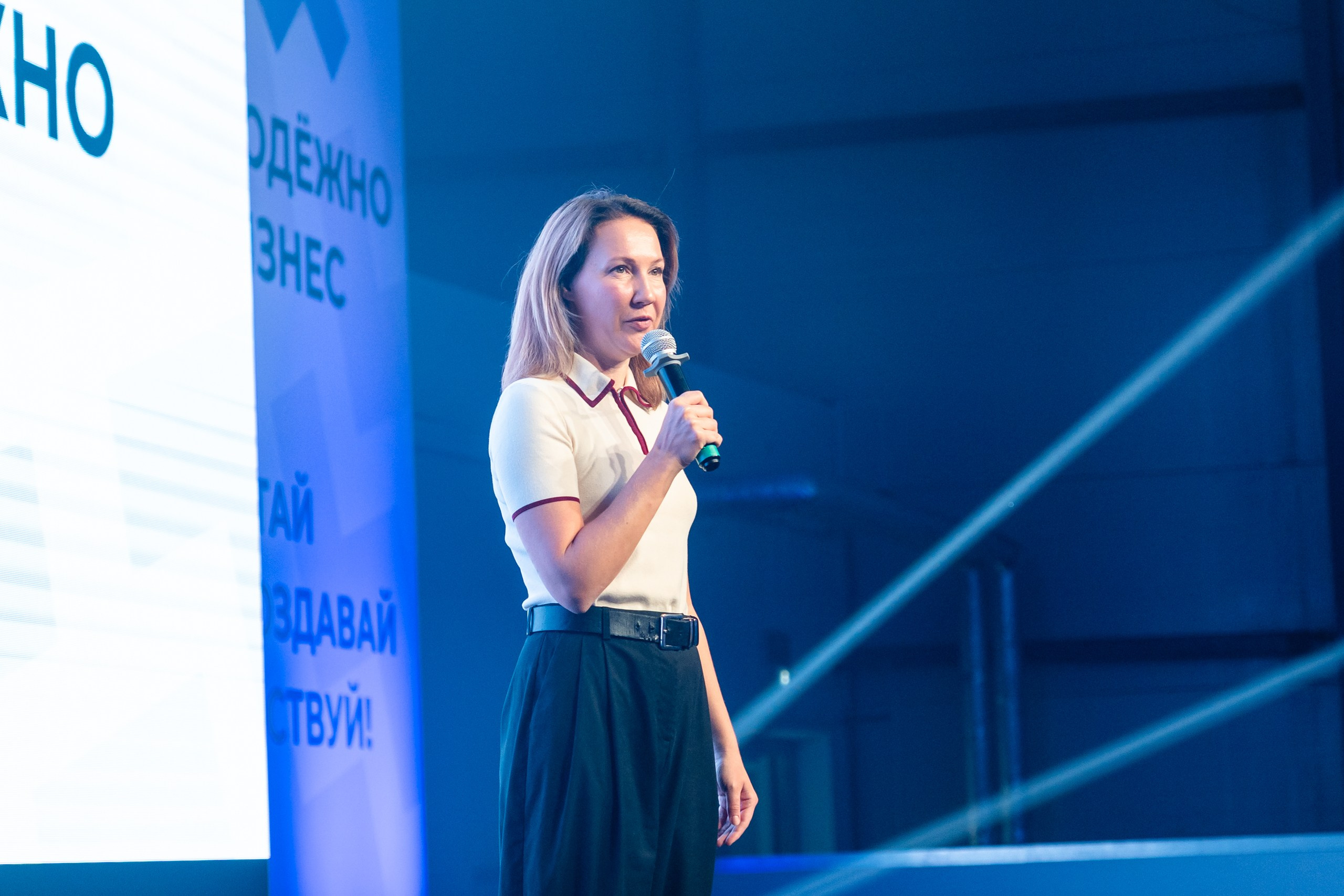 Репортажная съёмка форума: женщина-спикер с микрофоном выступает на сцене, синее освещение и баннеры «Молодёжный бизнес» на фоне
