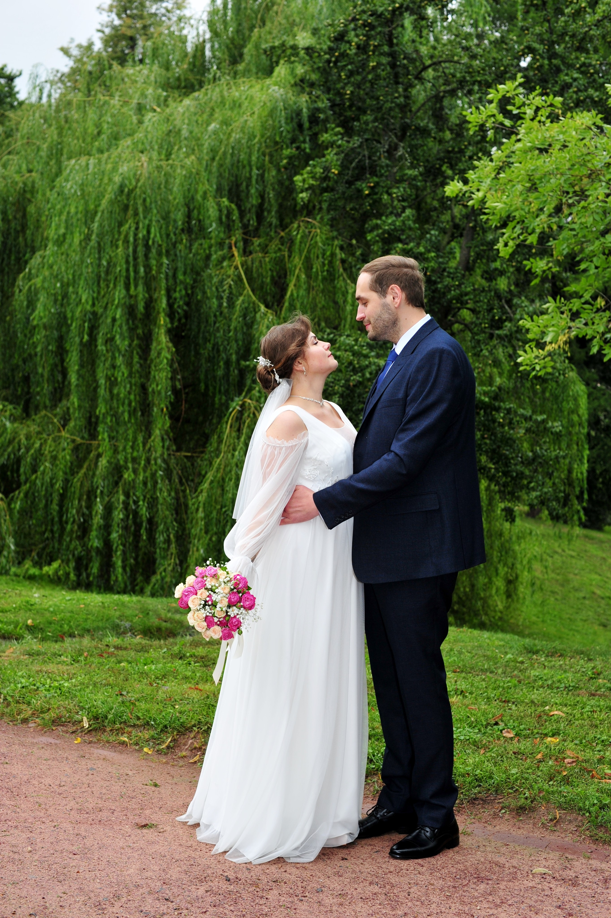 Свадьба в Царицыно пасмурным летним днем, новобрачные стоят на дорожке в парке и смотрят друг на друга на фоне зеленых деревьев