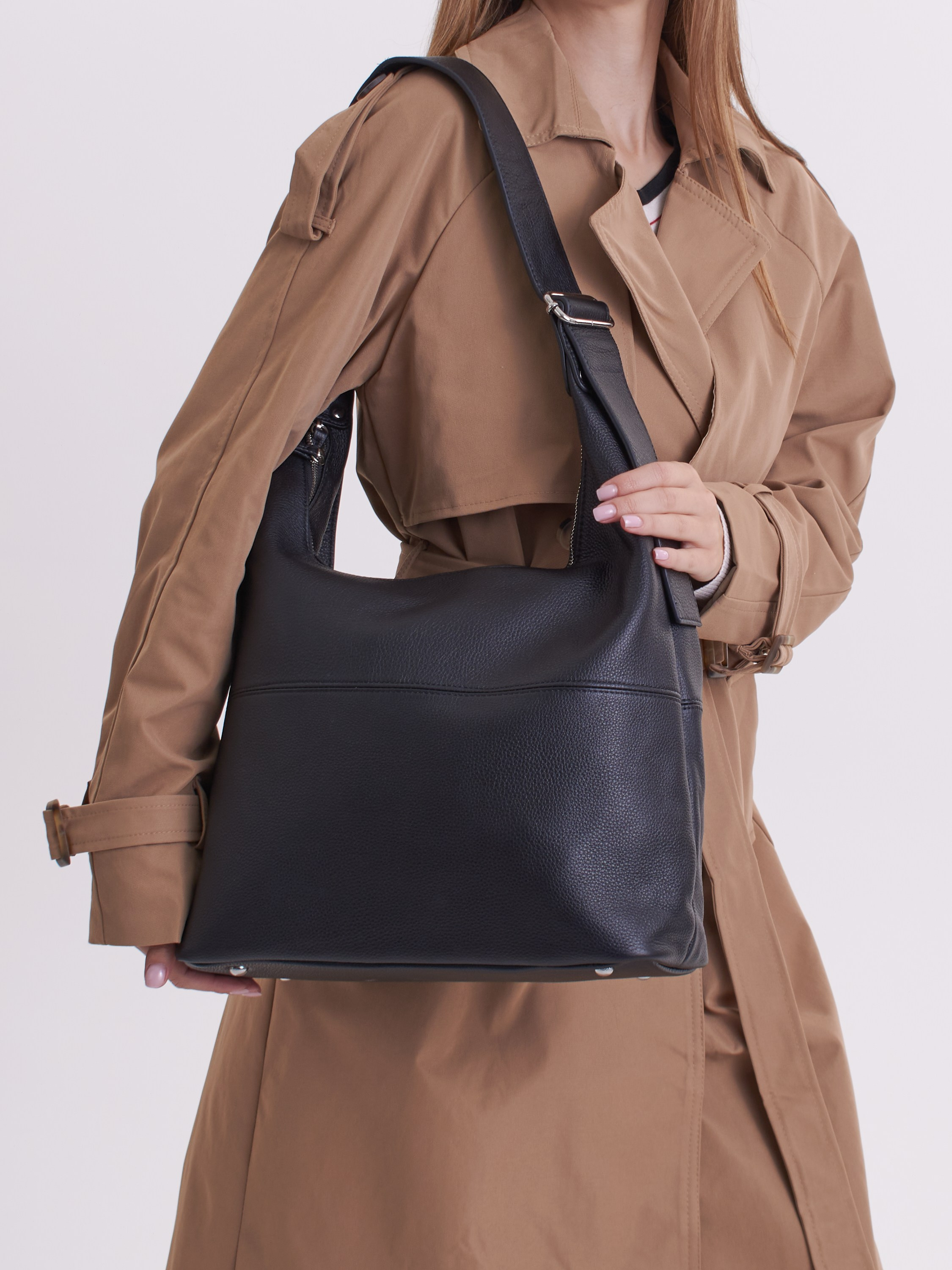 Девушка в плаще на светлом фоне держит на плече черную сумку 